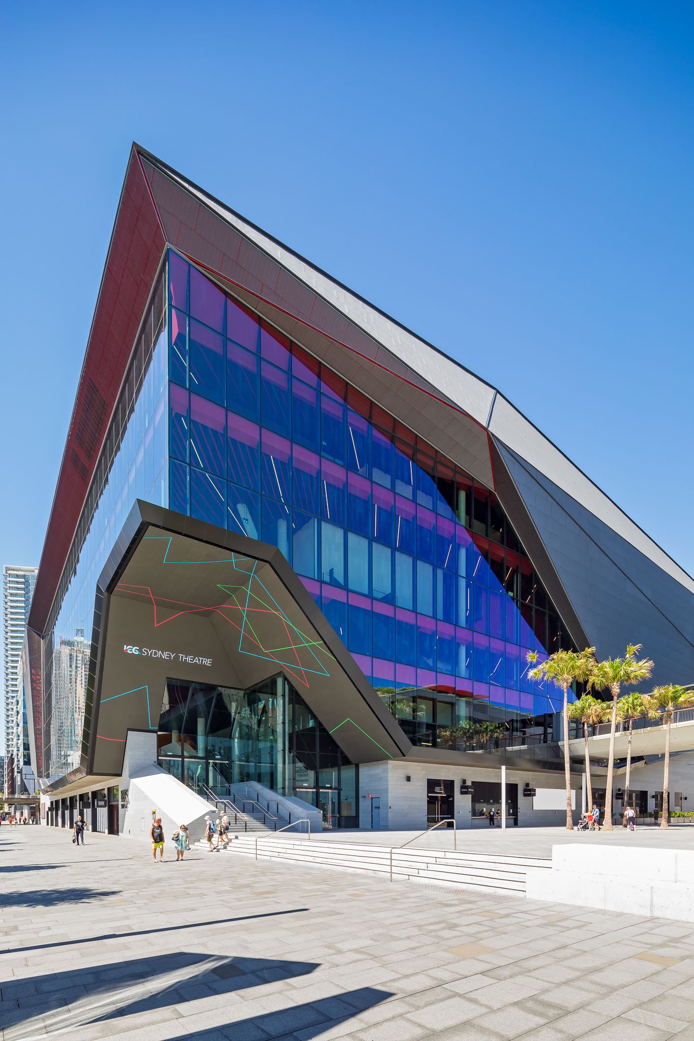 Dramatic angular theatre building with a reflective blue and pink glass facade, palm trees and a wide paved forecourt in the sunshine.