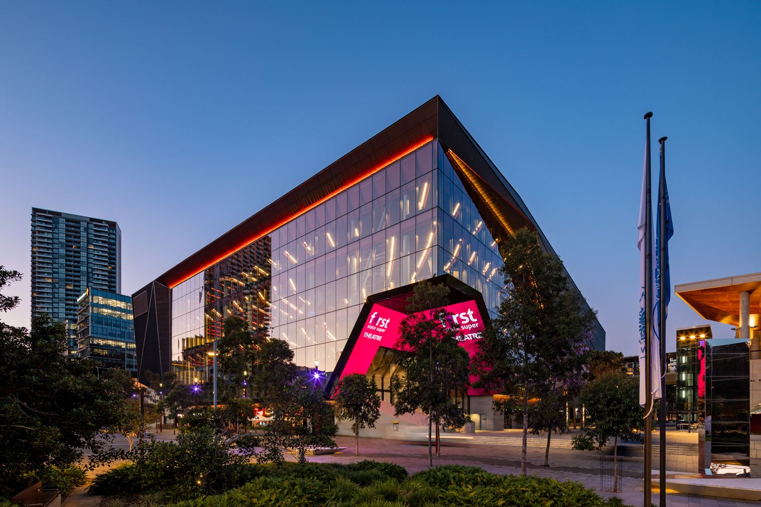 Angular glass convention building lit with red and pink lights at dusk beside tall city towers.