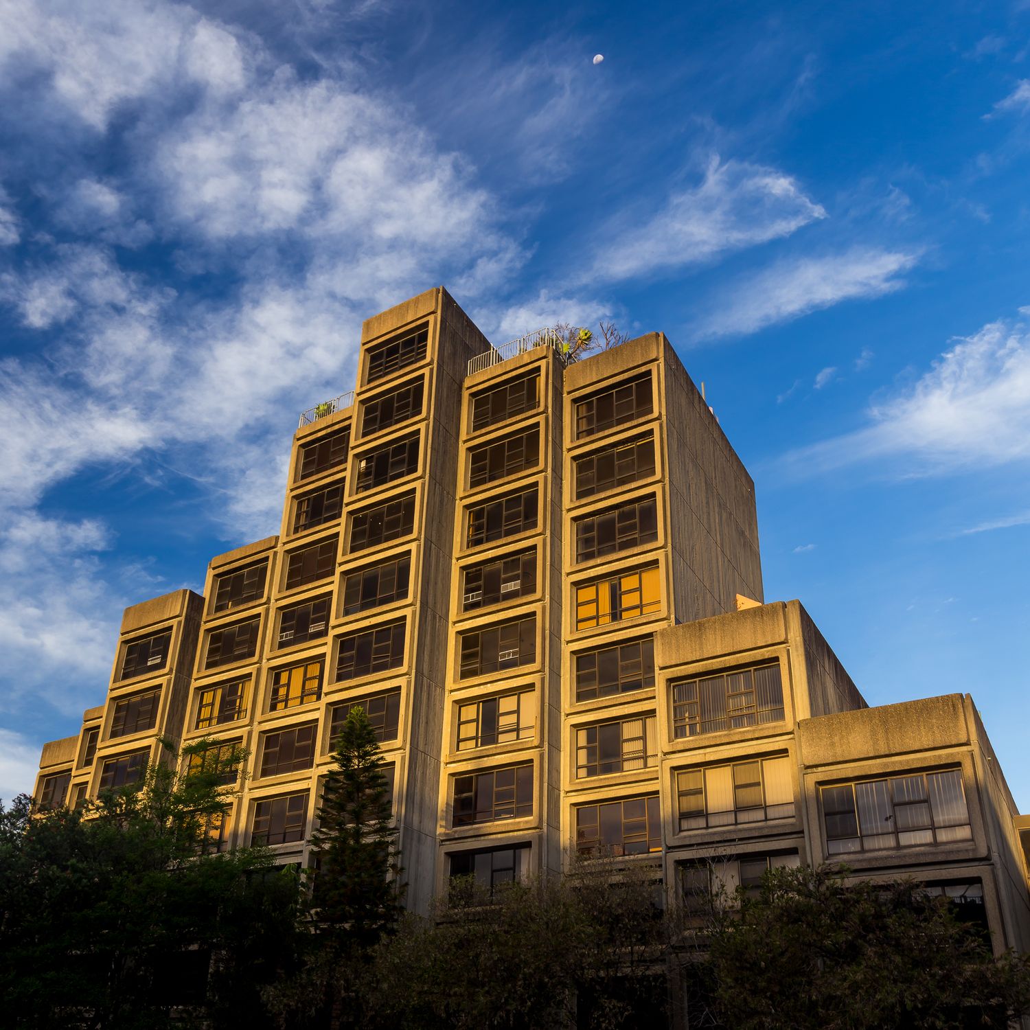 Brutalist stepped apartment building of tan concrete with rows of recessed windows under a blue sky with wispy clouds.