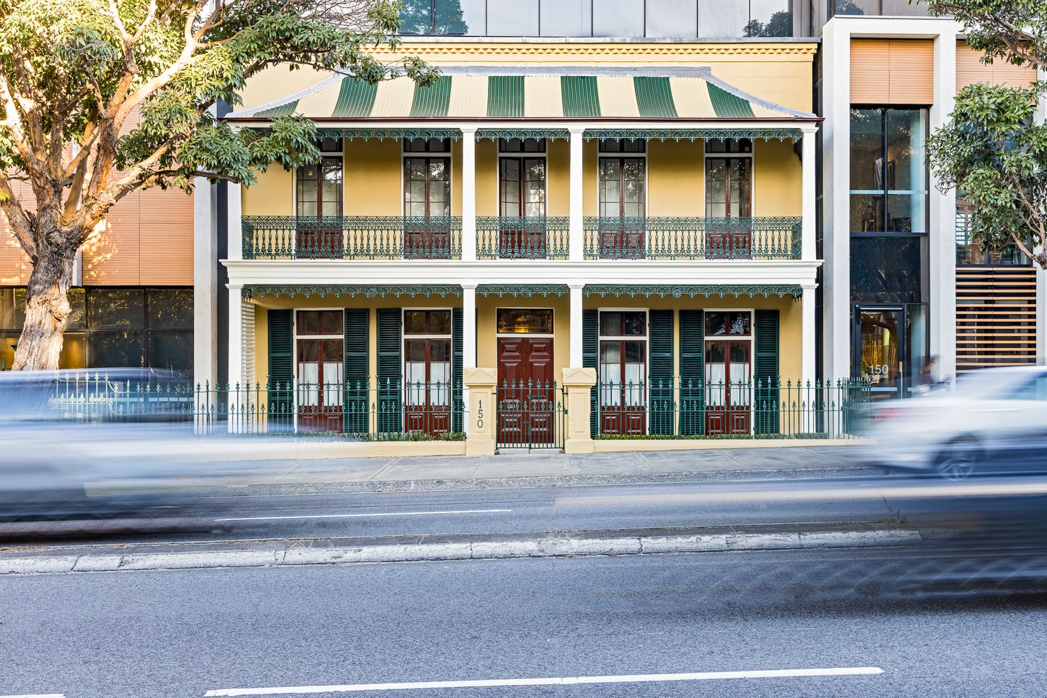 Historic two-storey terrace building with green shutters, iron lacework balcony and red timber doors beside a busy city street.