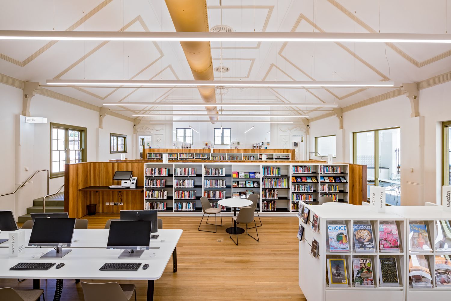 Sunlit library interior with timber floors, tall bookshelves, study tables with computers and a vaulted ceiling with exposed beams.