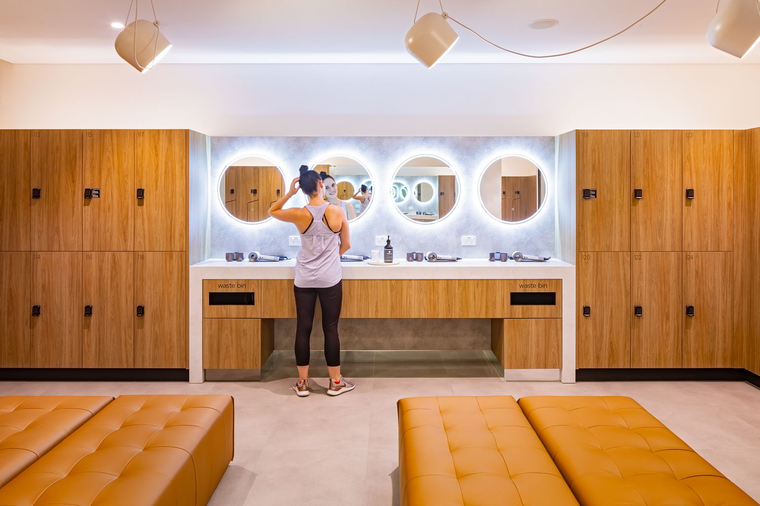 Modern locker room with warm wood lockers, round backlit mirrors and tan leather benches as a woman fixes her hair.