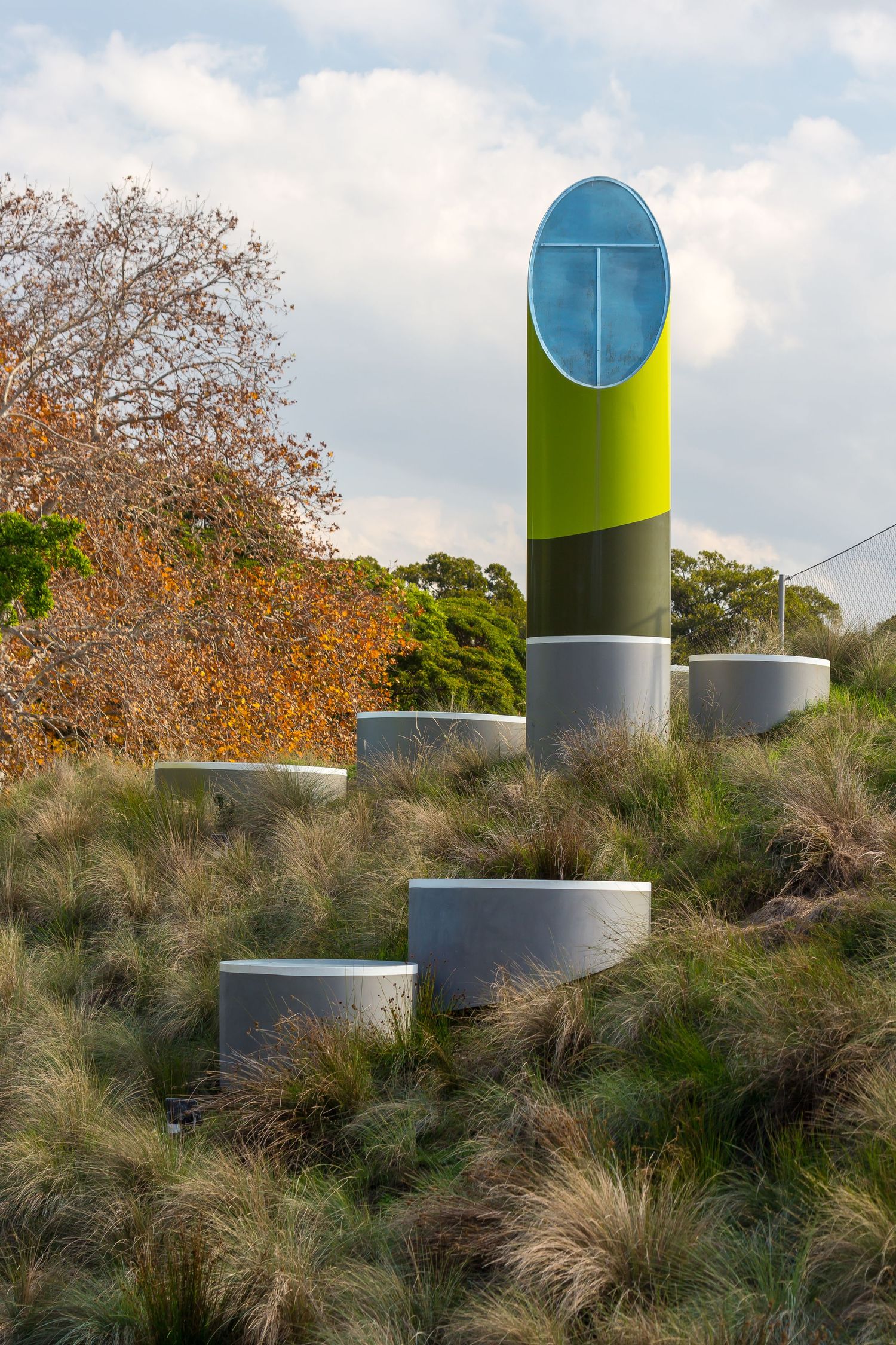 Tall lime green and gray cylindrical sculpture with a blue circular top rising above round concrete seats in a grassy landscaped park.