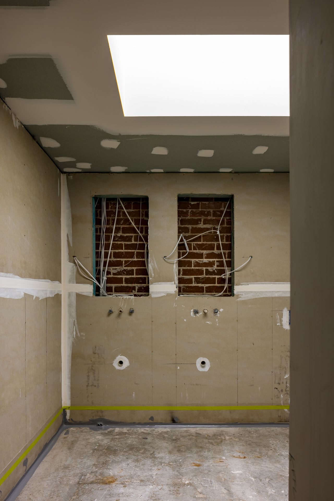 Bathroom under construction with exposed brick wall recesses, loose wiring, plumbing outlets and a bright skylight overhead.