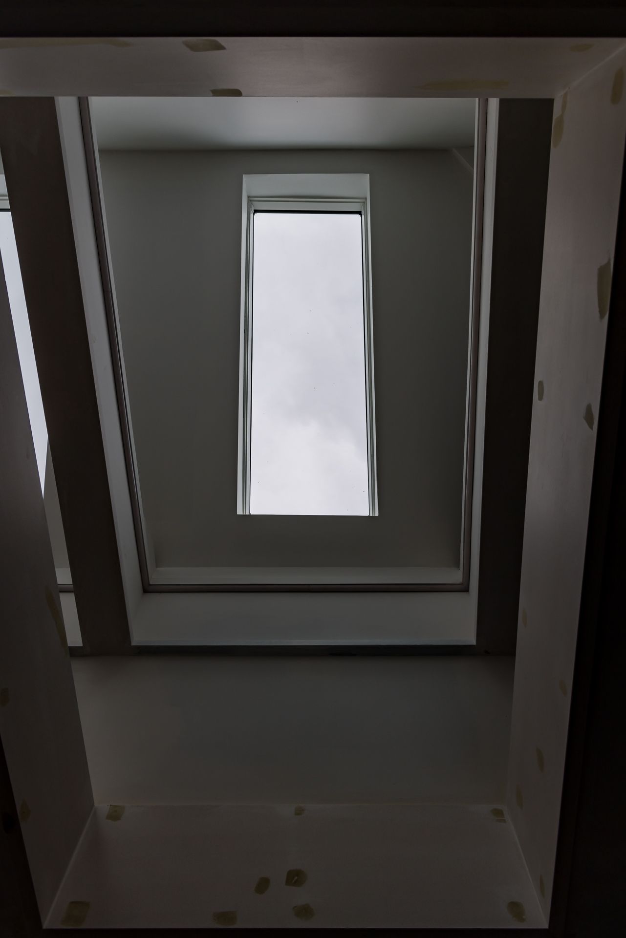 Upward view of a deep skylight shaft with a tall narrow window showing a cloudy grey sky.