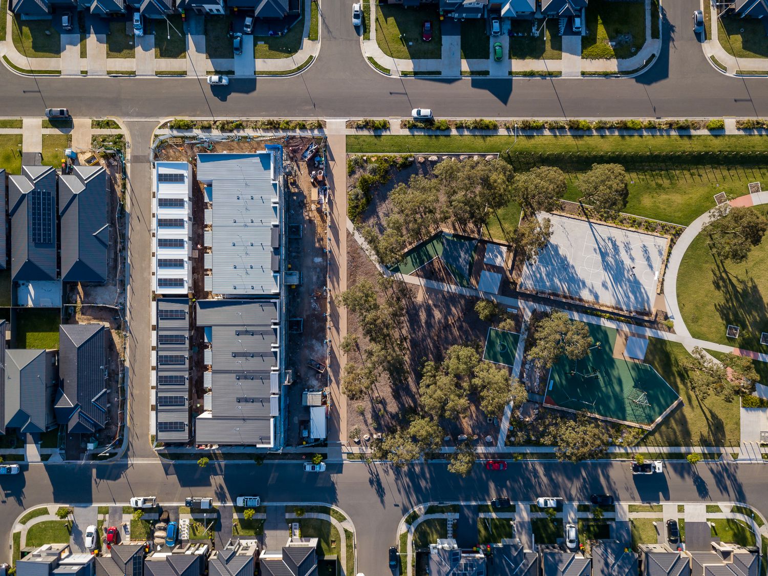 Aerial view of new townhouses and a long apartment block beside a park with tennis courts and tree-lined streets.