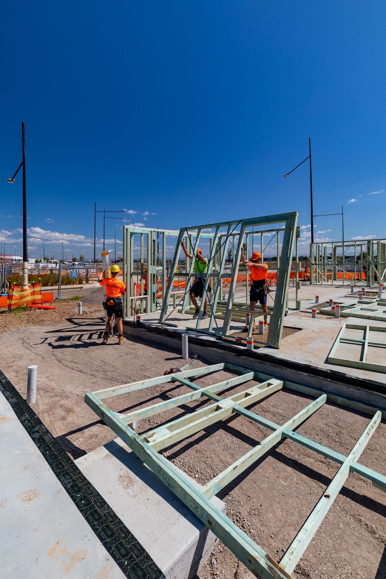 Construction workers in hi-vis gear assemble green steel wall frames on a concrete slab under a clear blue sky.