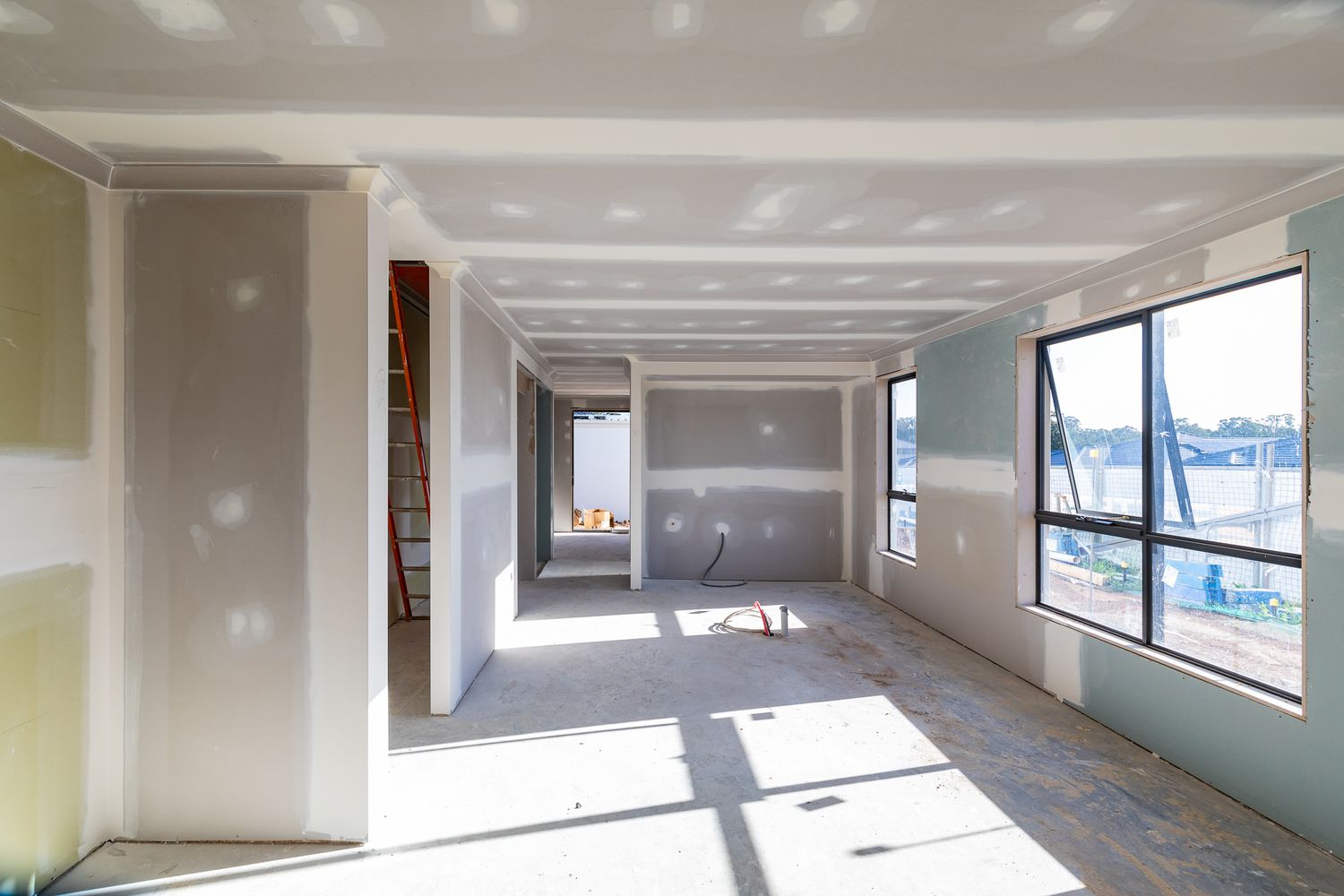 Bright interior of a new home under construction with plasterboard walls, large windows and sunlight on the concrete floor.