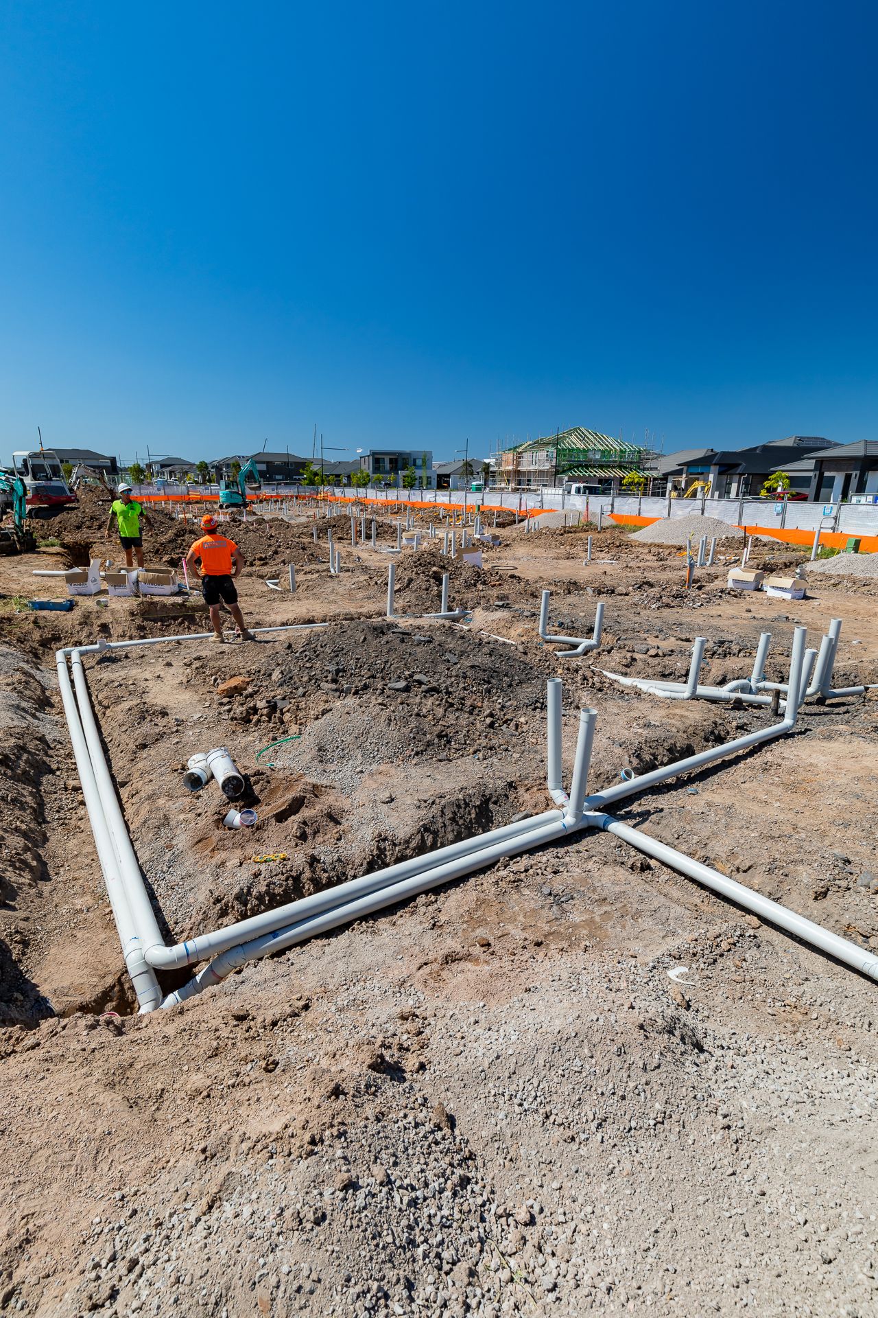 Workers inspect grey plumbing pipes laid in trenches across a dirt building site before house slab construction begins.