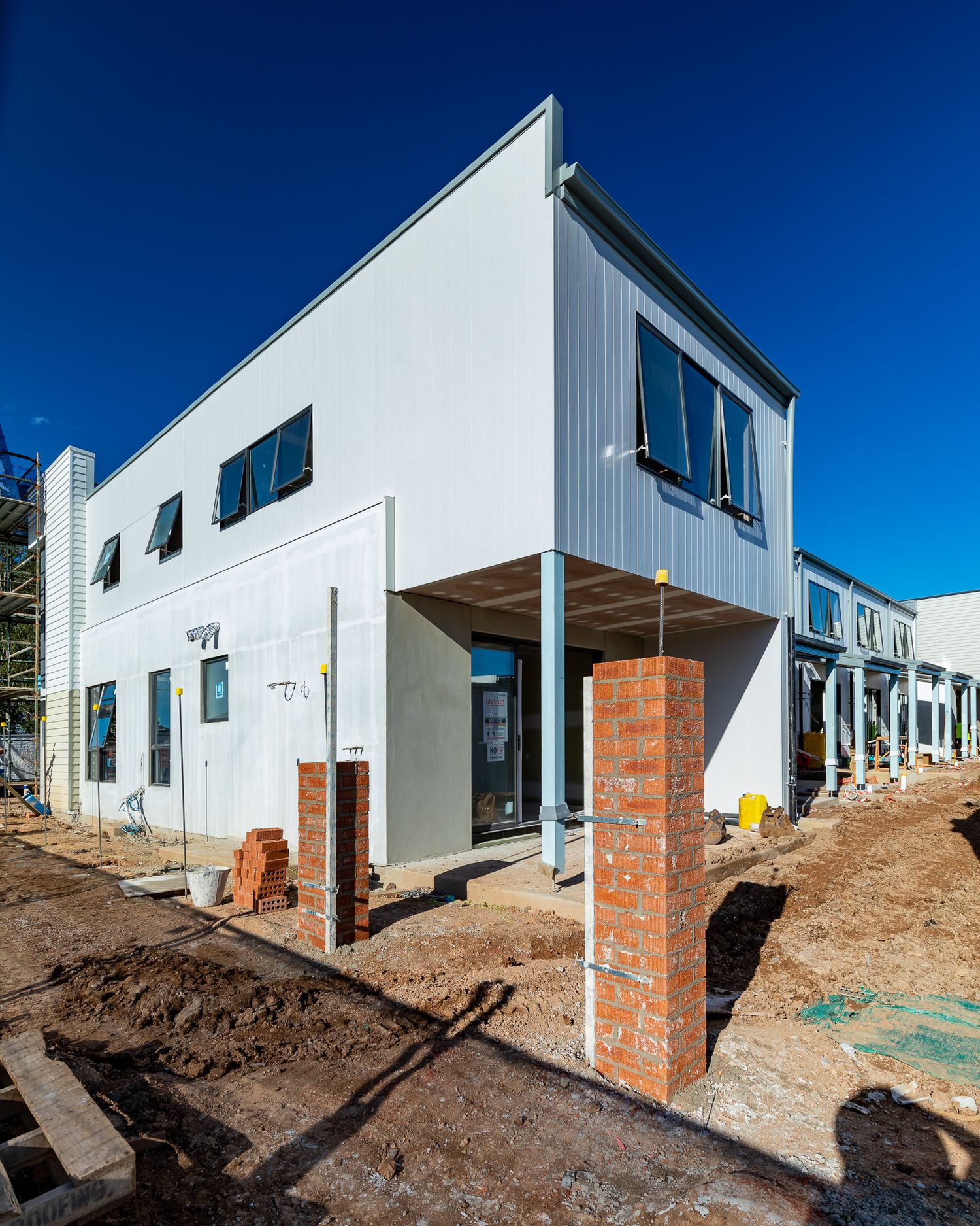 New two-storey townhouse with white metal cladding and a brick pillar under construction on a dirt site beneath a clear blue sky.
