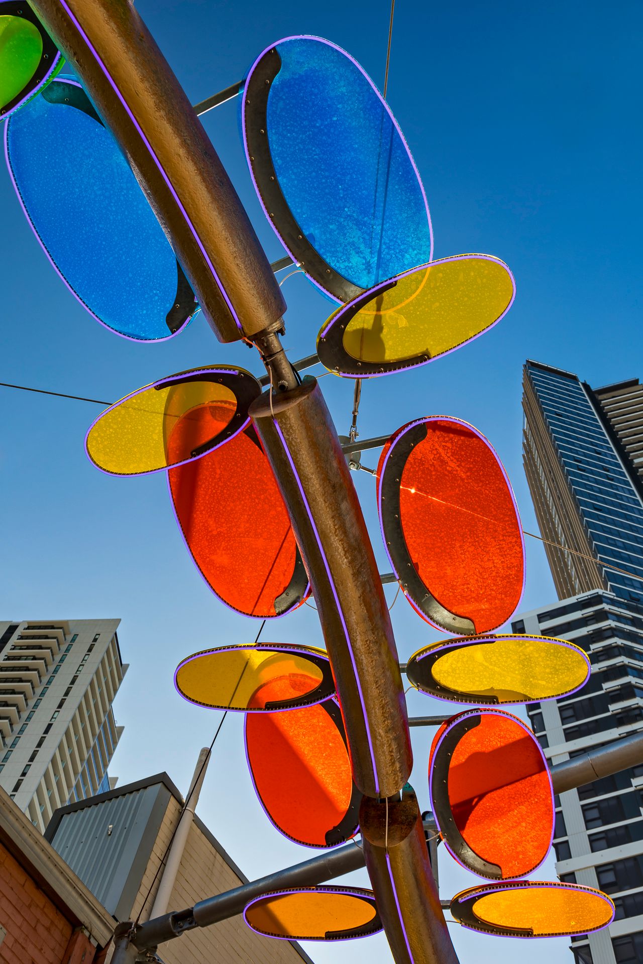 Close view of translucent blue, yellow and orange discs on a curved bronze sculpture with city towers behind.