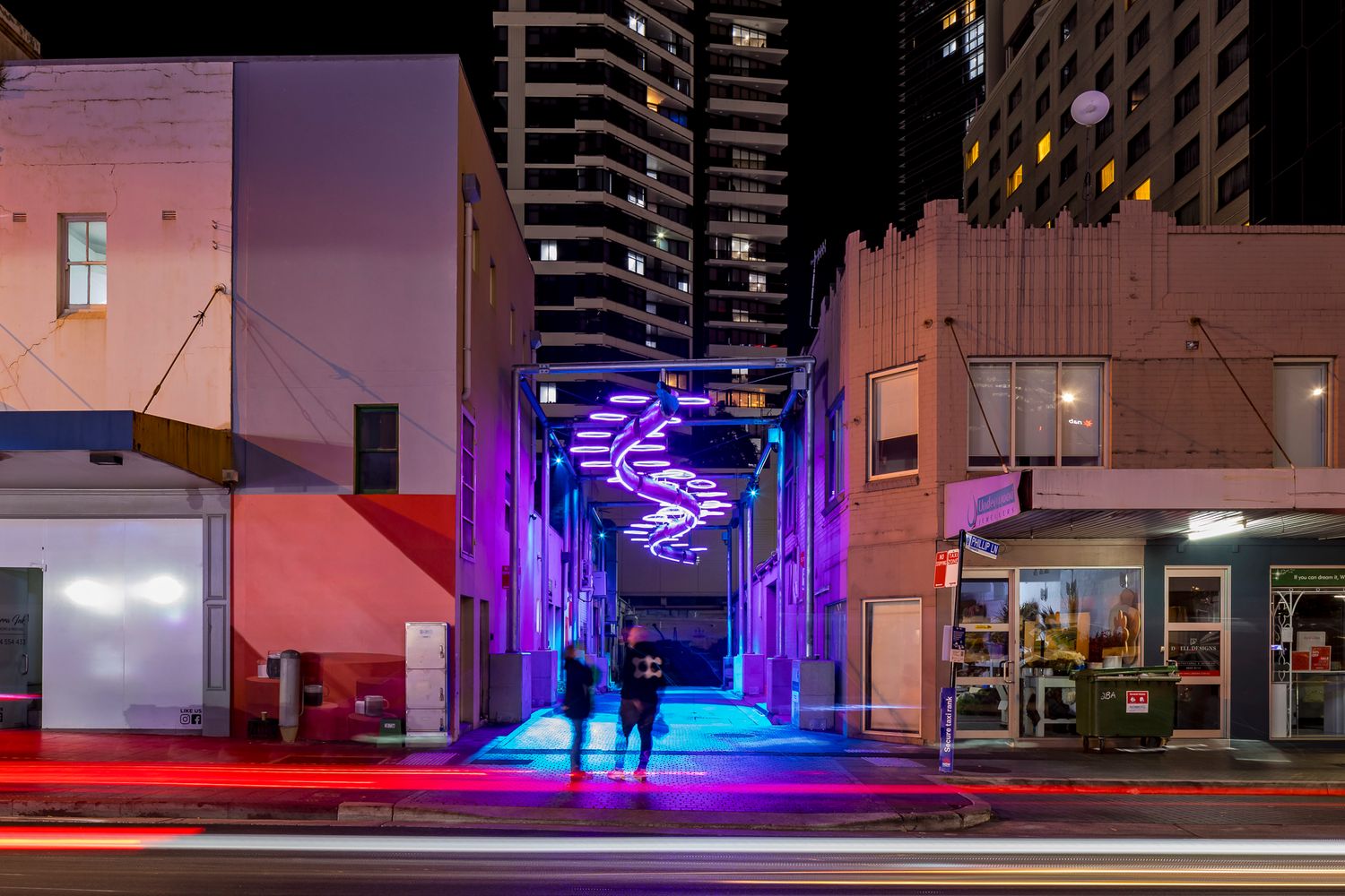 Pink and violet neon spiral installation lights up a city laneway at night with long-exposure light trails on the street.