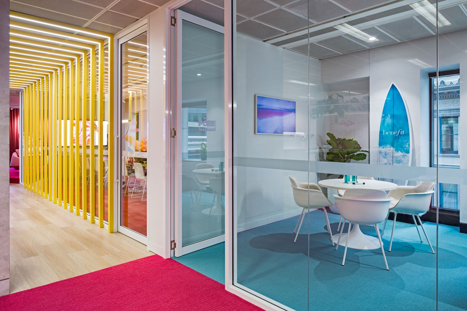 Colorful office interior with pink carpet, yellow slatted partition, glass walls and a white round table with chairs on teal flooring.