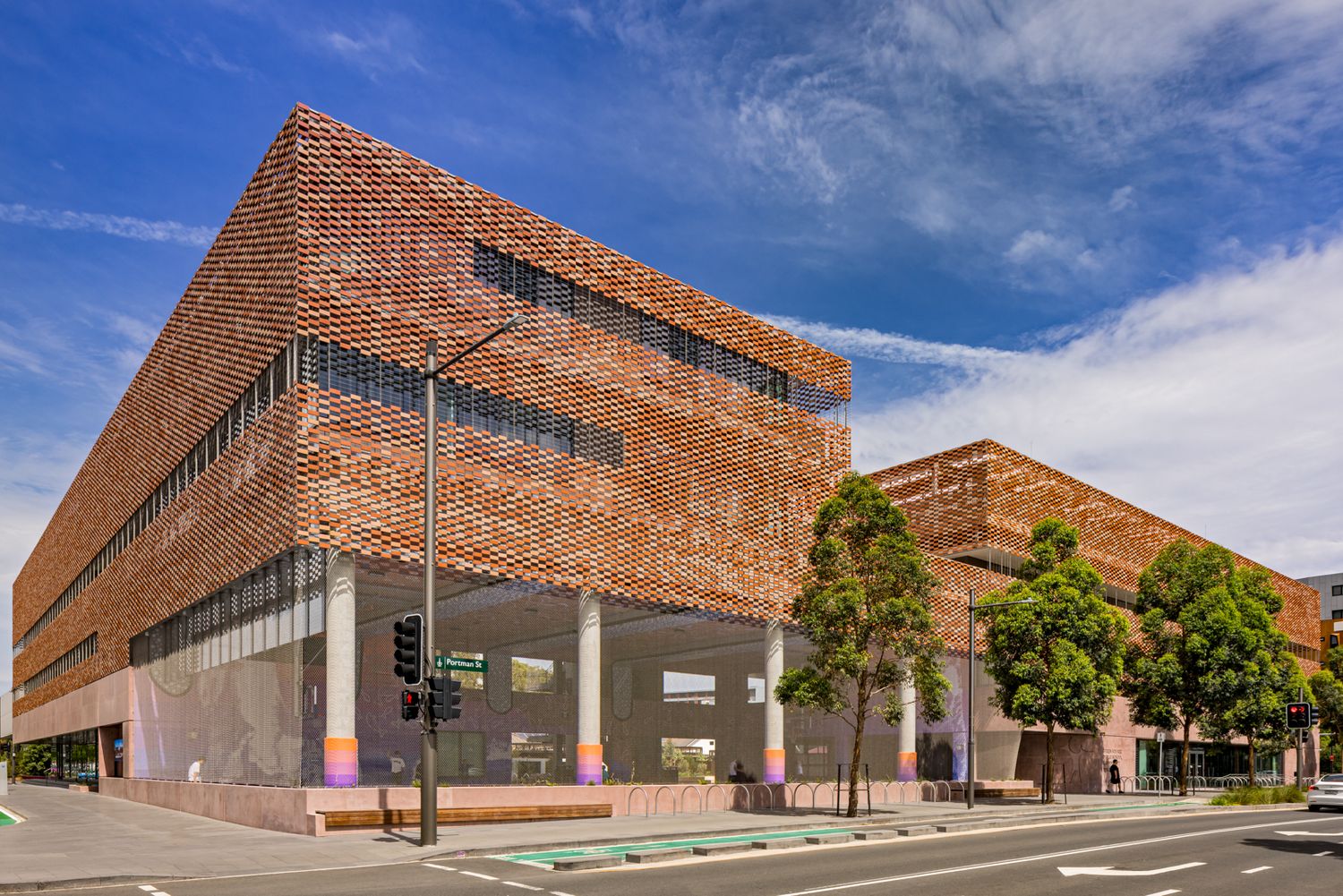 Contemporary building with a textured perforated terracotta brick facade and white columns beside a tree-lined city street.