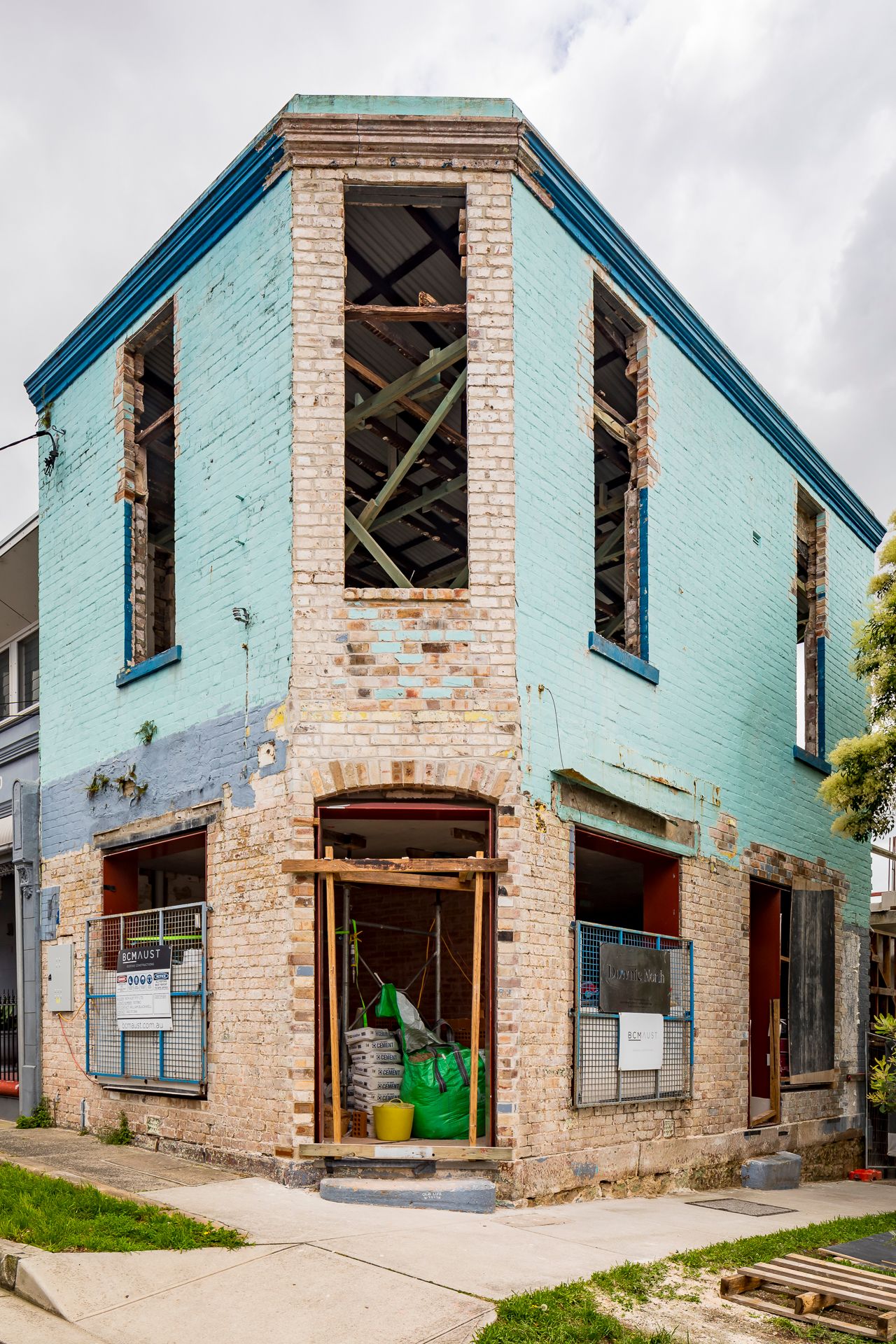 Two-storey corner brick building under renovation with turquoise painted walls, exposed timber framing and open window frames.