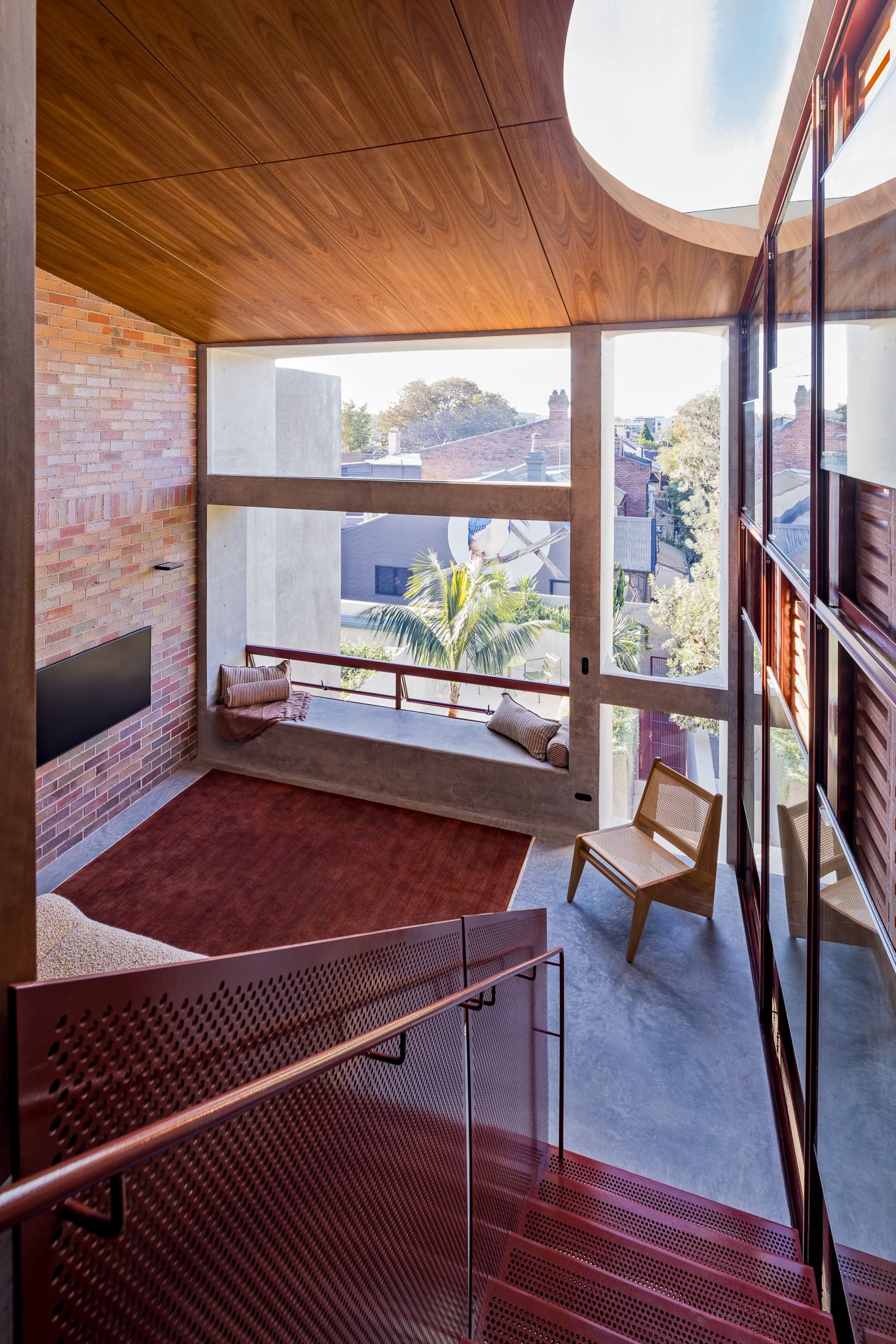 Upper-floor lounge with a window seat, red rug, wall-mounted television, timber ceiling, skylight, and a red perforated stair balustrade.