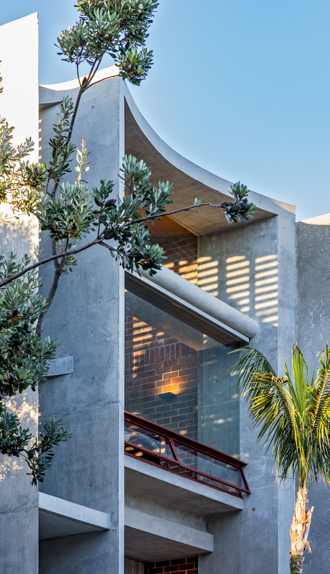 Curved concrete townhouse facade with a red balustrade and glowing interior brick wall, framed by a native tree and palm frond.