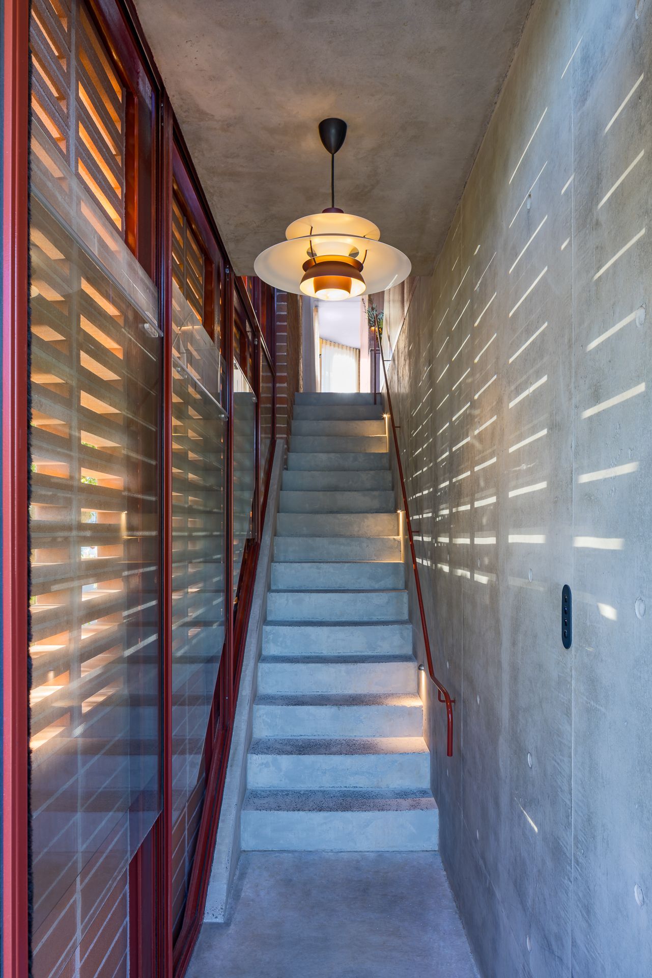 Narrow concrete staircase with red handrail and vintage pendant light, lit by sunlight streaming through timber louvres.