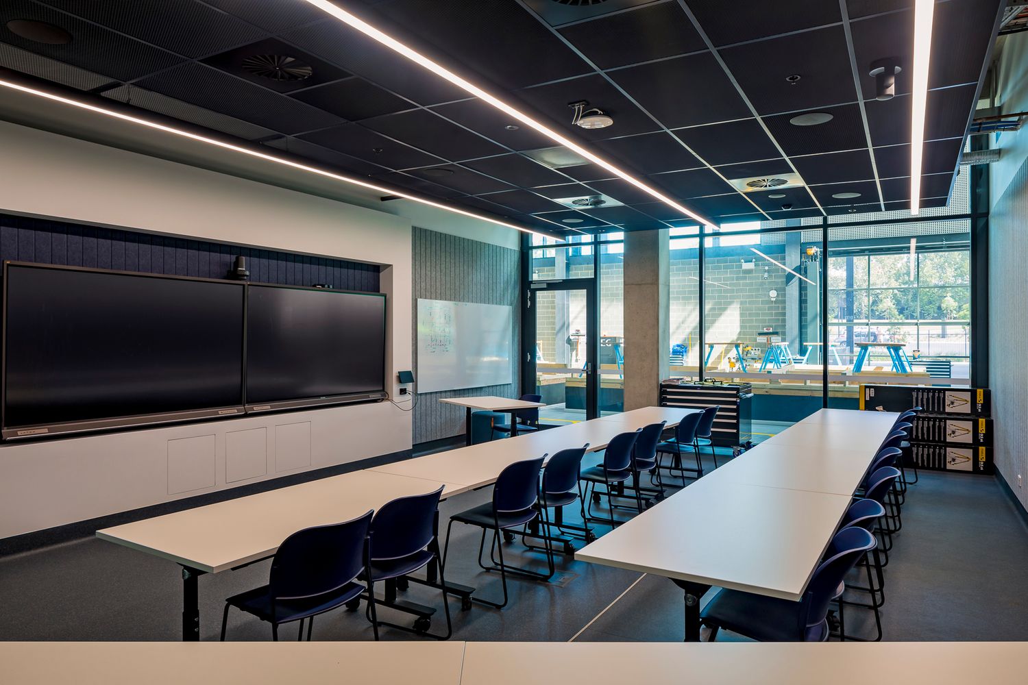 Modern TAFE classroom with long white tables, navy chairs, a wall-mounted display screen and glass walls overlooking a bright study area.