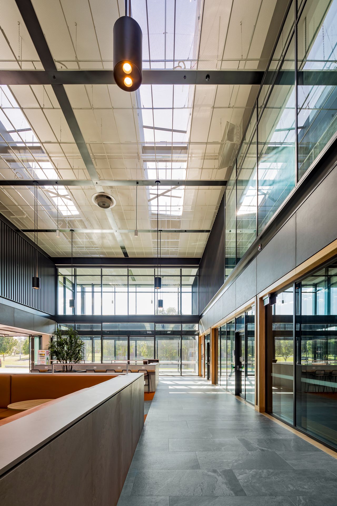 Light-filled TAFE atrium with exposed steel beams, skylights, pendant lighting, timber-framed glazing and polished stone flooring.