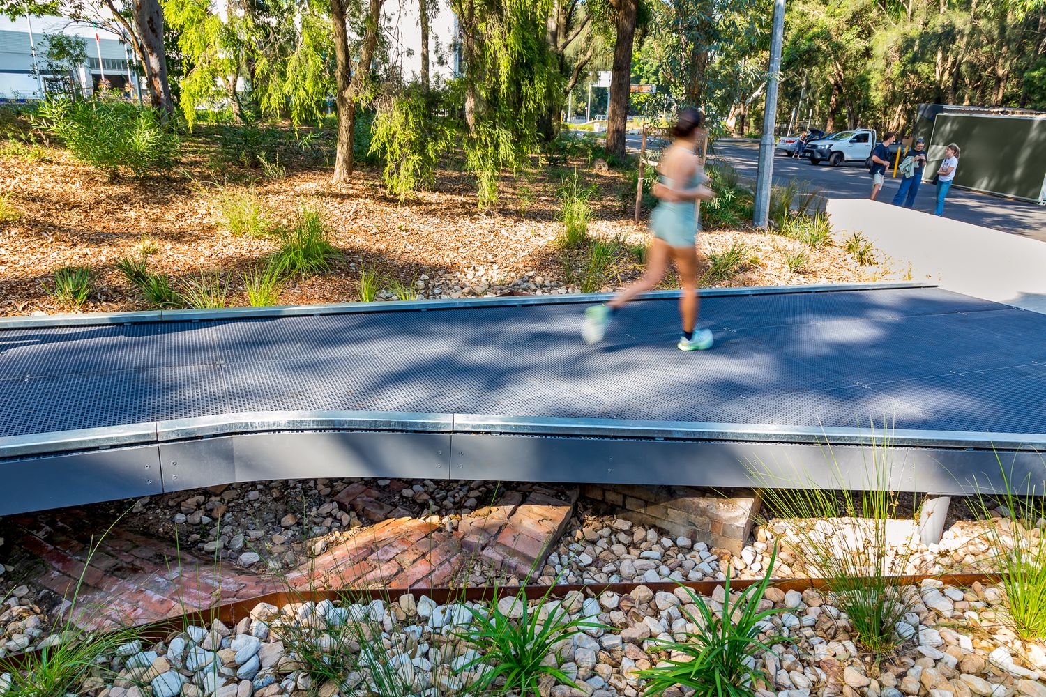 A jogger runs across a curved dark footbridge above river pebbles and native plantings in a leafy urban park.