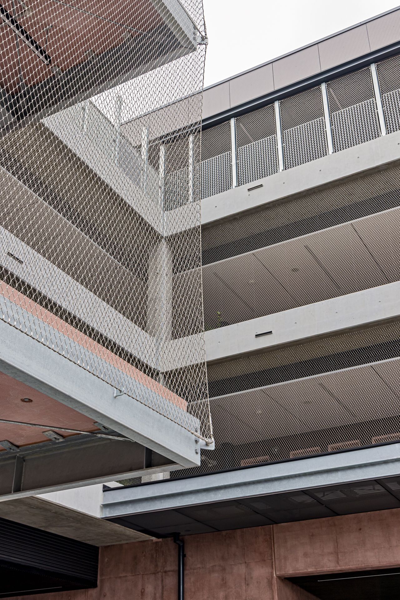 Modern multi-level parking structure with open concrete floors, metal mesh safety netting, and glass-paneled railings under a bright sky.