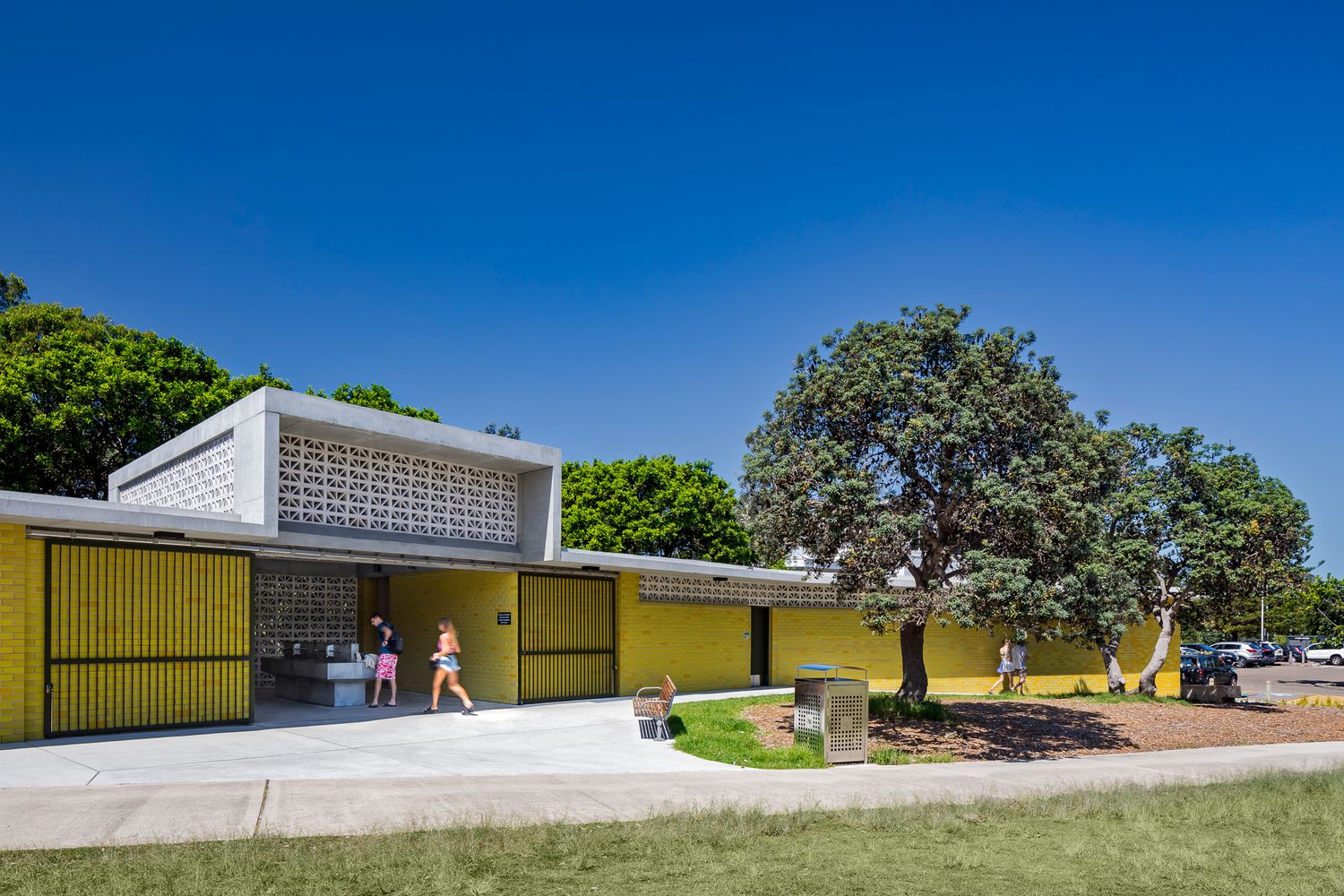 Visitors stand near the open entrance of a mid-century style yellow brick public amenities building surrounded by trees and lawn.