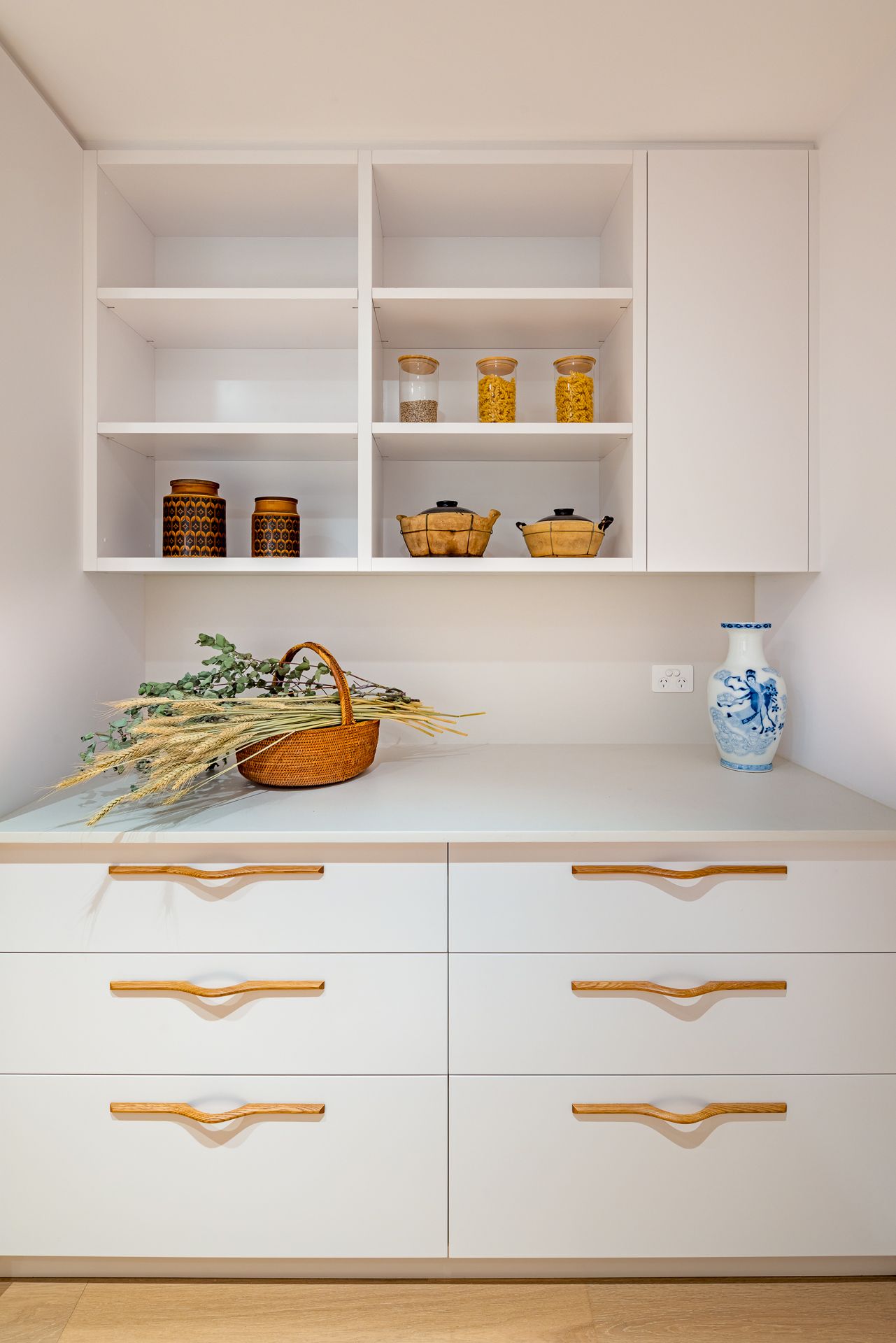 White built-in cabinetry with open shelves, timber handles, glass jars, a woven basket of dried foliage and a blue-and-white vase.