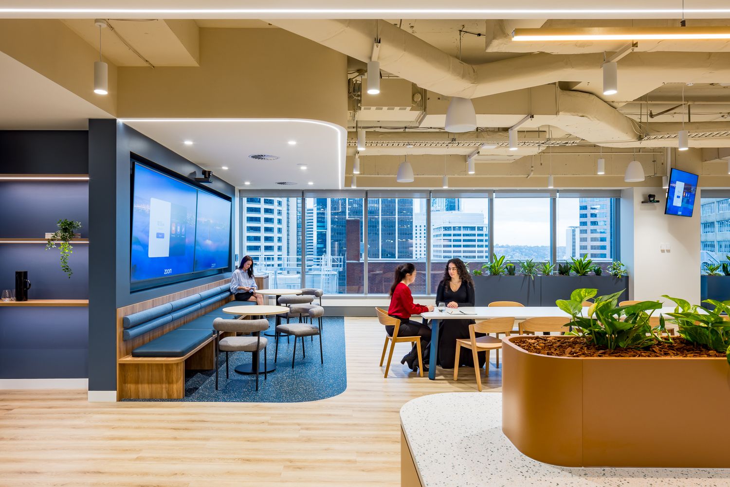Two colleagues chatting at a table in a bright modern office lounge with banquette seating, greenery and skyline views.