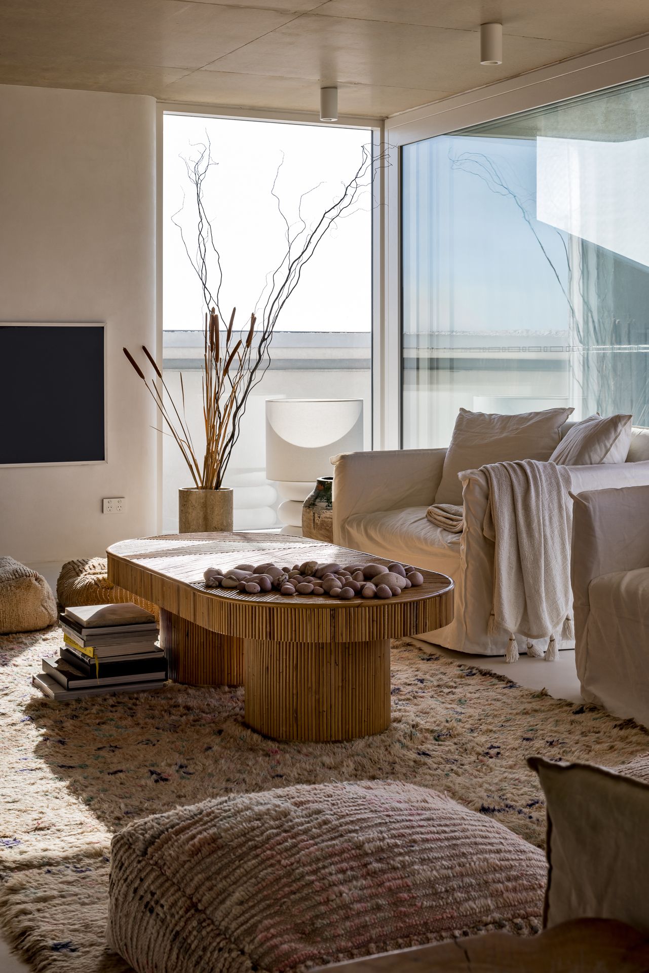 Cozy neutral living room with a round wood coffee table, white sofa, woven rug and tall dried branches beside large windows.