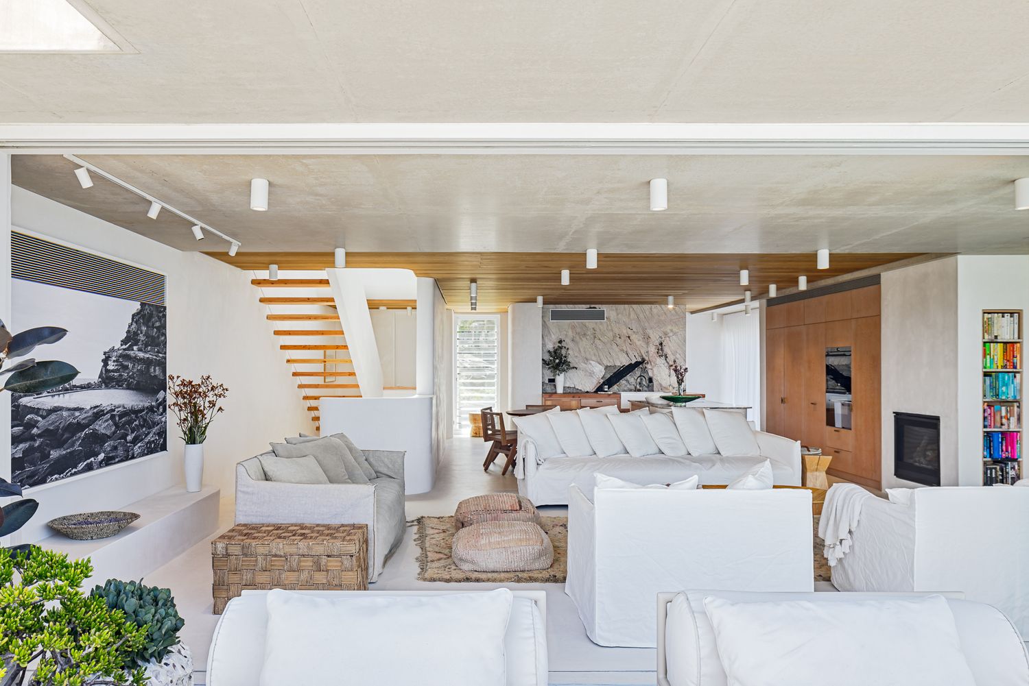 Spacious coastal living room with white sofas, timber staircase, wood-slat ceiling, artwork and a view toward the kitchen.