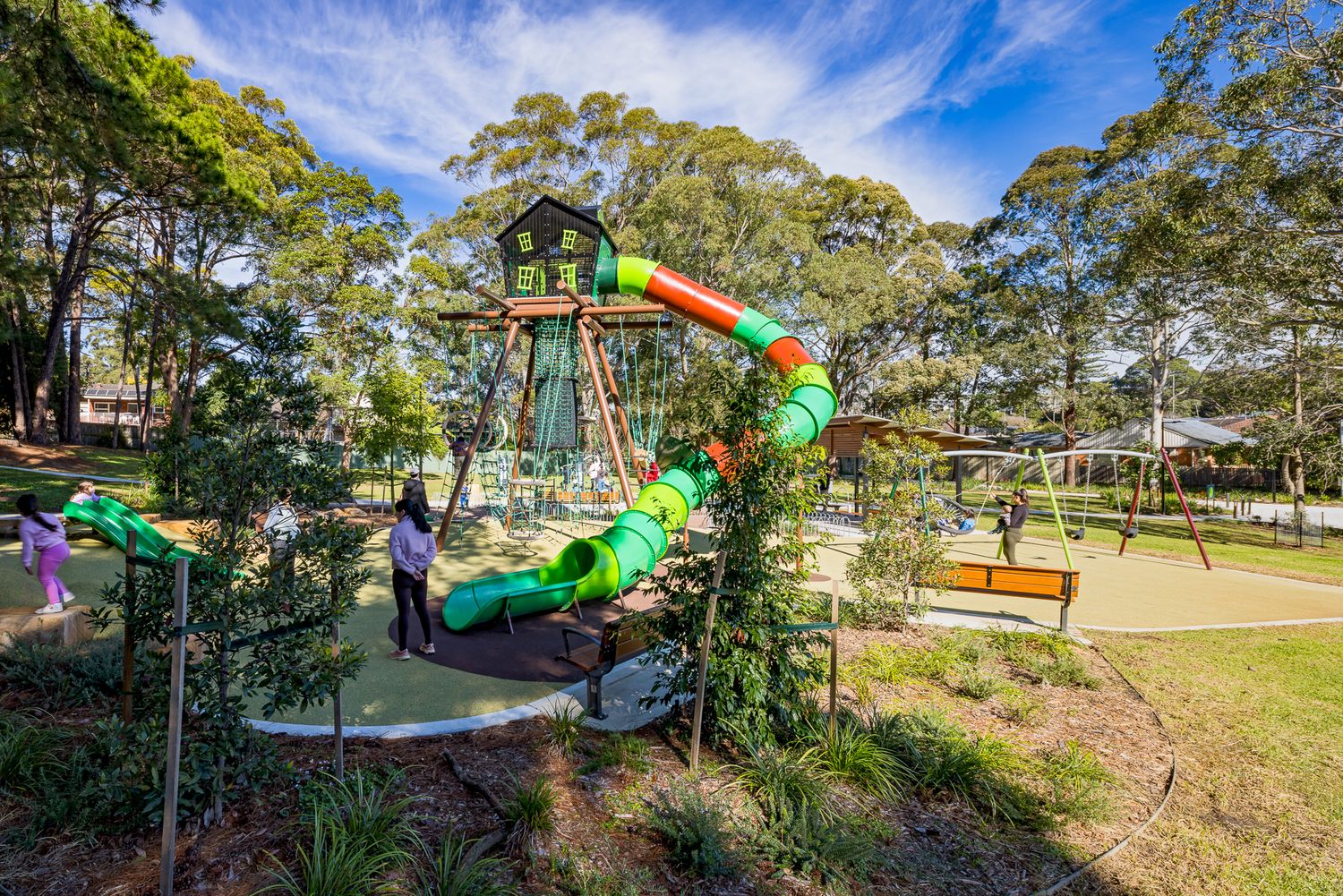 Children play on a treehouse-style playground with a long spiral tube slide, surrounded by eucalyptus trees and soft green lawn.