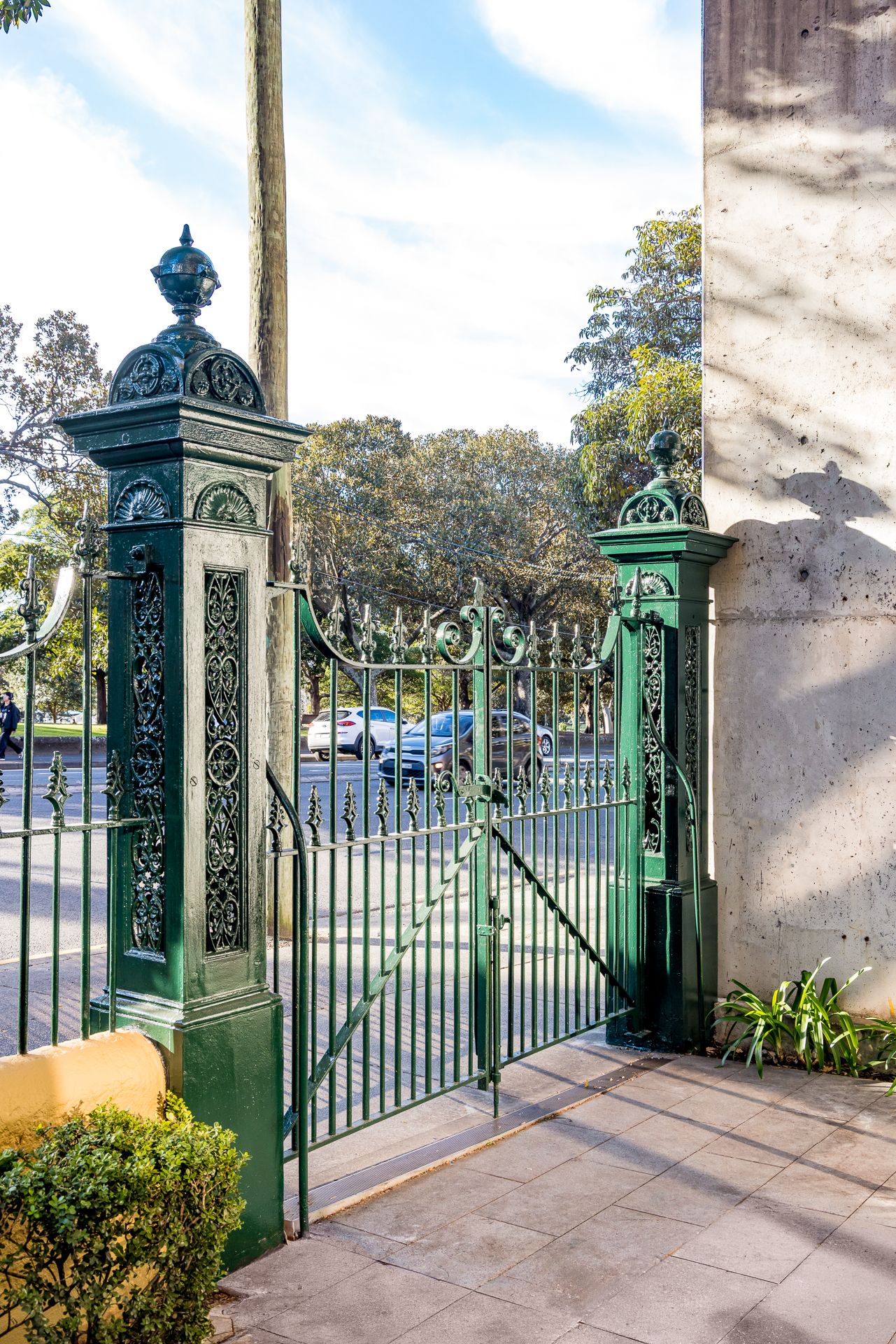 Ornate green cast-iron gate and decorative gate posts opening onto a tree-lined park and street.