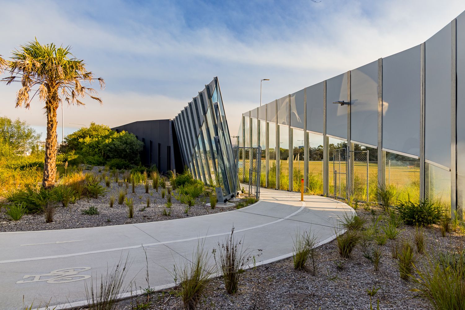 Curving concrete path beside a modern angled glass and metal-panel building, surrounded by palms, gravel garden and native grasses.