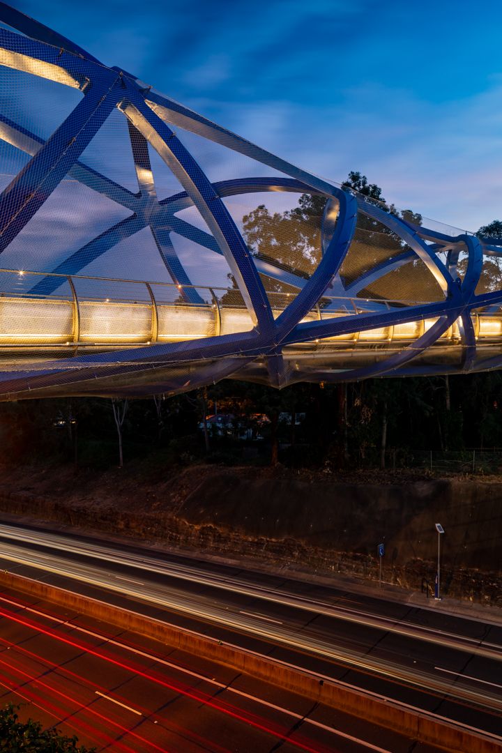 Curved blue steel pedestrian bridge glowing at twilight over a busy motorway, with red and white light trails streaking below.