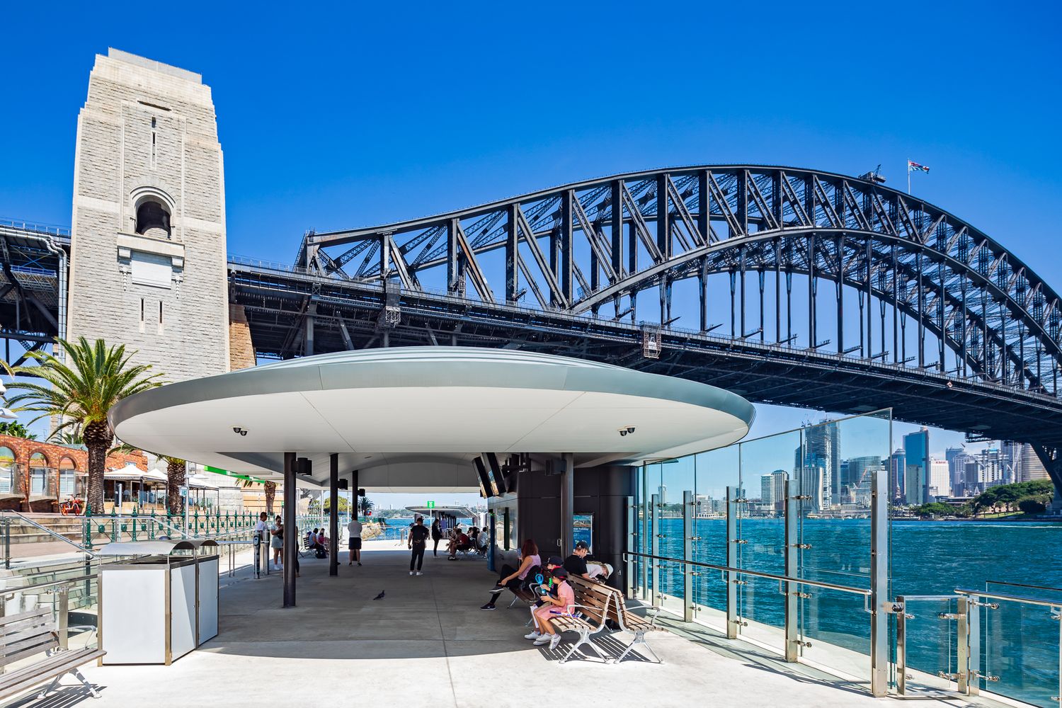 Ferry wharf shelter with an oval roof beneath Sydney Harbour Bridge, where passengers relax on seats beside the sparkling harbour.