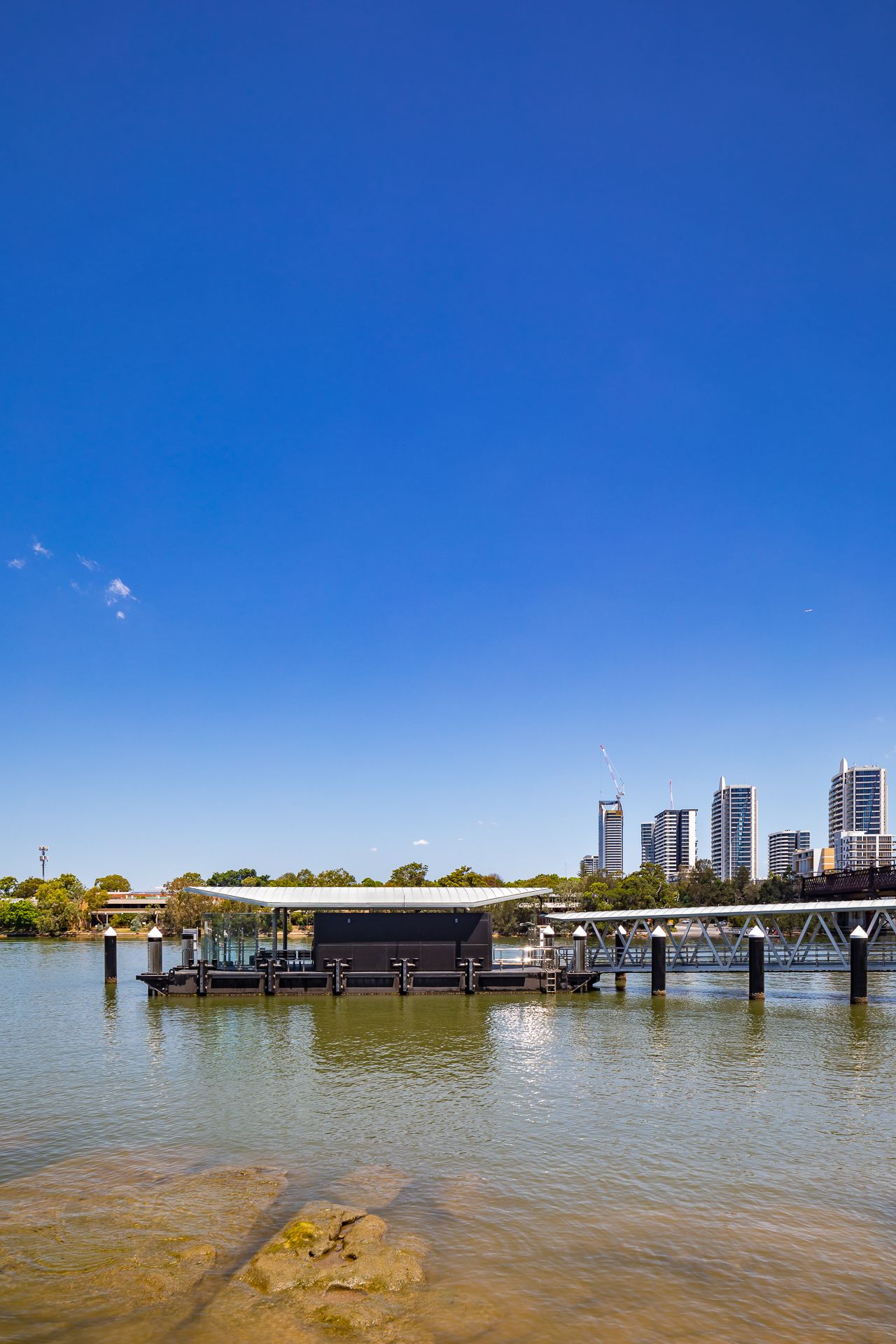 Floating ferry pontoon with a low canopy on a calm river, with city high-rise buildings in the distance under a deep blue sky.
