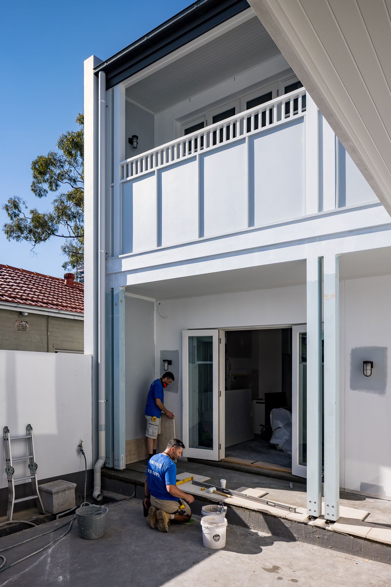 Two tradesmen work on the rear patio of a two-storey white house with a balcony and open French doors during a home renovation.
