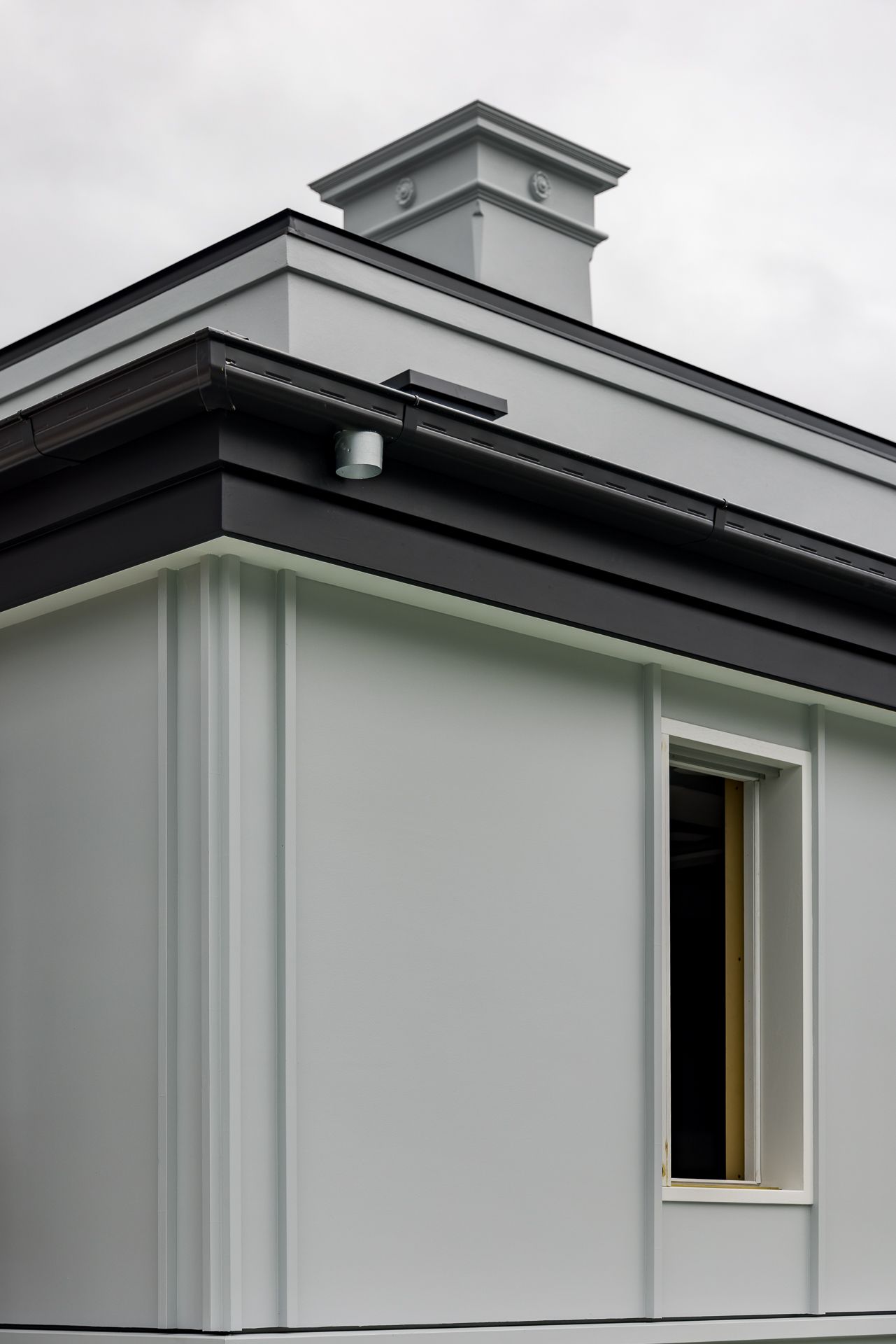 Exterior corner of a modern grey rendered building with dark gutter trim, a small window and a decorative chimney under an overcast sky.