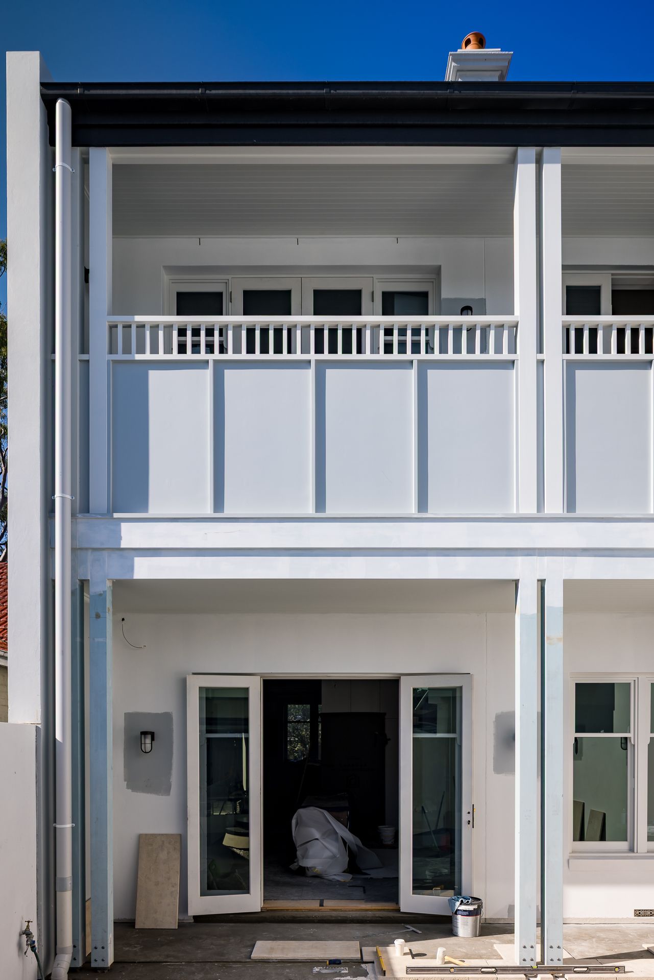 White two-storey townhouse rear with a balustrade balcony, French doors and construction materials near the ground floor entrance.