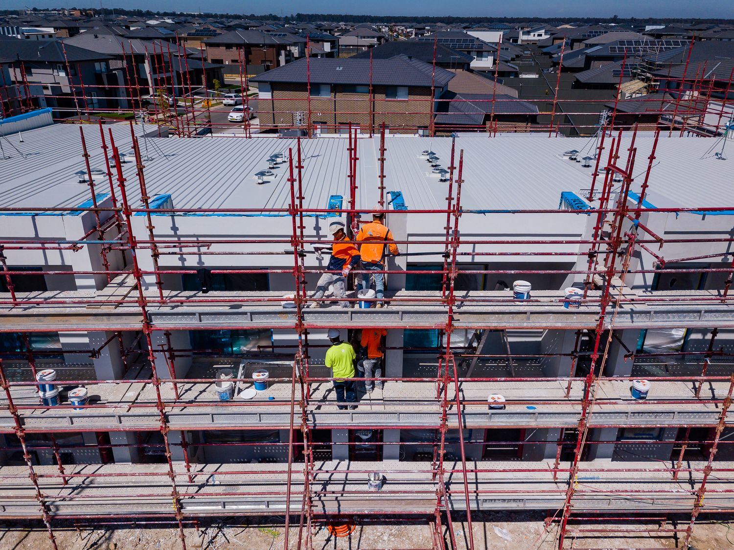 Construction workers in high-visibility clothing standing on scaffolding around a multi-storey townhouse building with a flat roof.