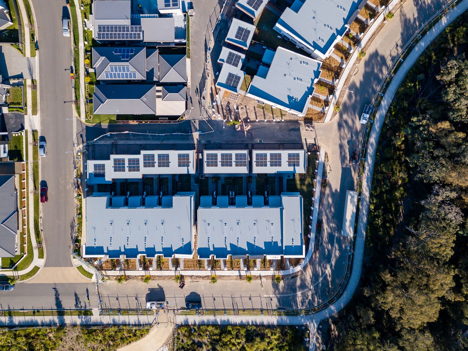 Overhead aerial view of modern townhouses with blue-grey rooftops beside a curved road and dense bushland.