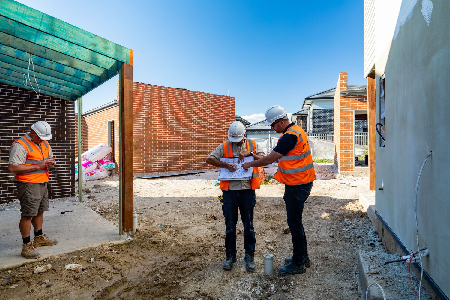 Three construction workers in hard hats and hi-vis vests discuss plans beside a new brick home with a covered entry.