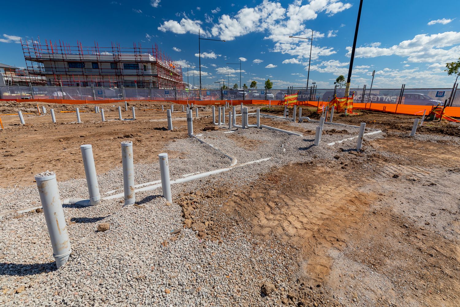 White drainage pipes rising from gravel at a construction site with a multi-storey building frame behind orange safety fencing.