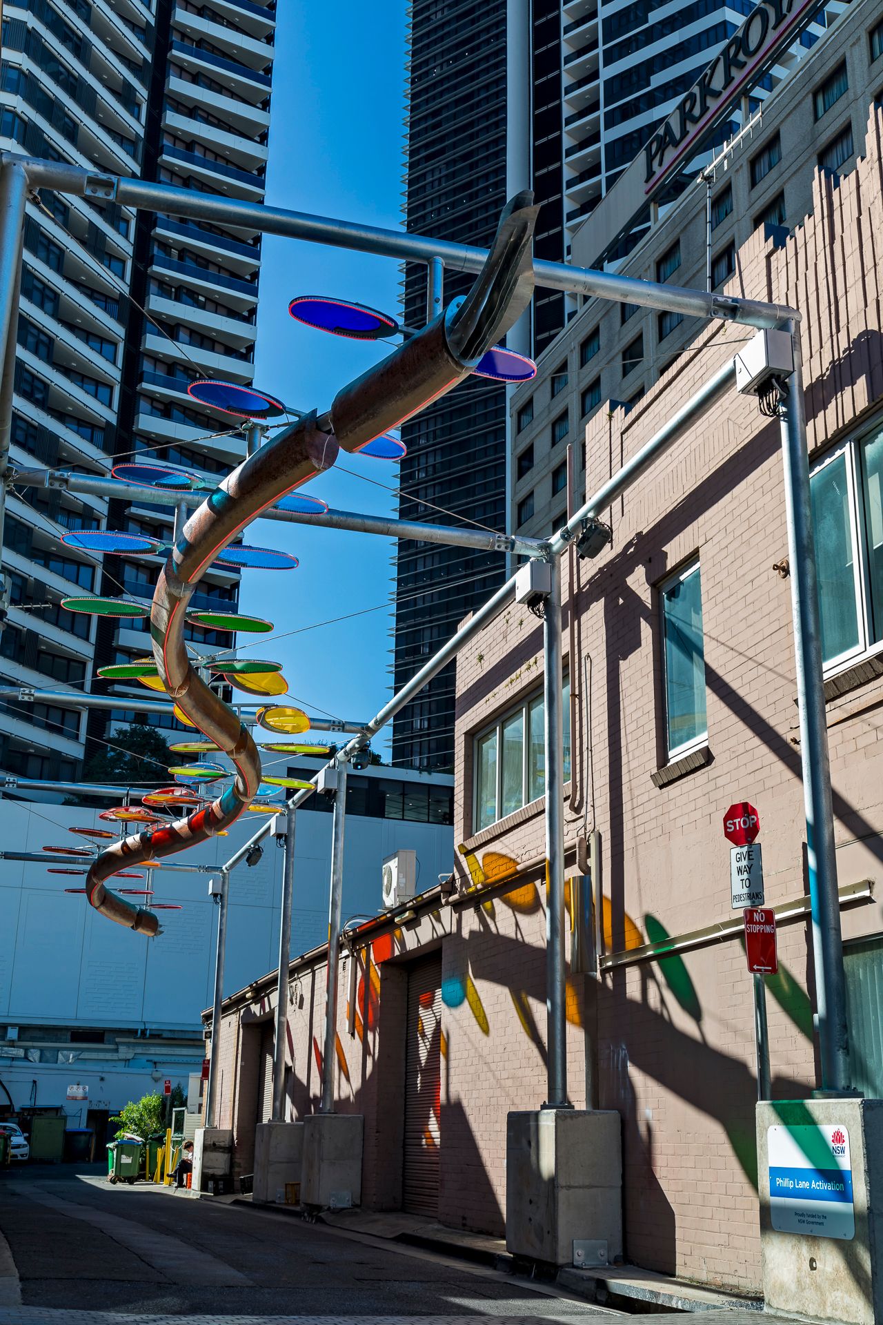 Colorful disc sculpture overhead in a sunlit urban alley, casting orange and green shadows on a pink brick wall.