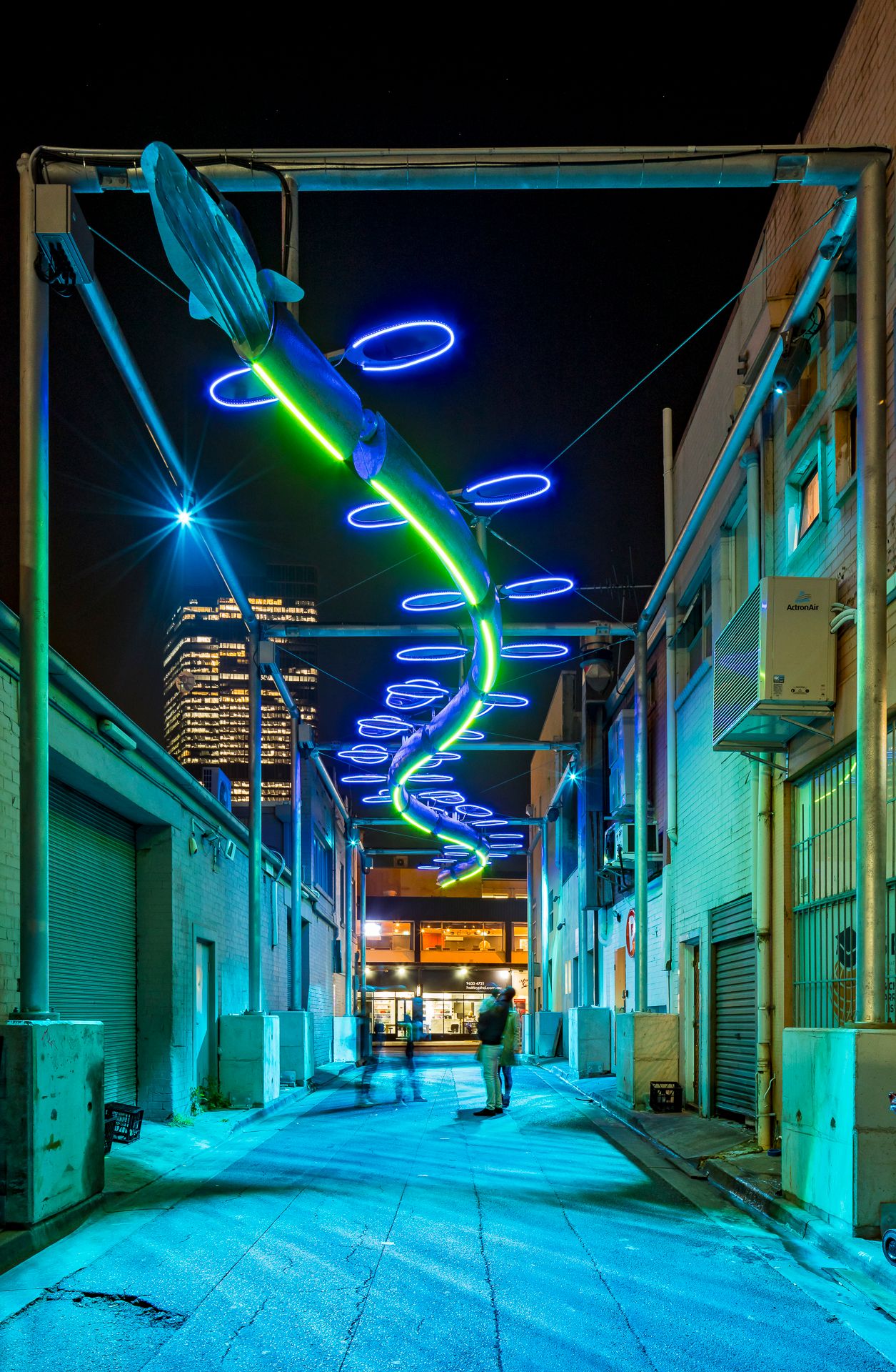 Two people stand in a quiet urban alley under a spiral light art installation glowing electric blue and green against the night sky.