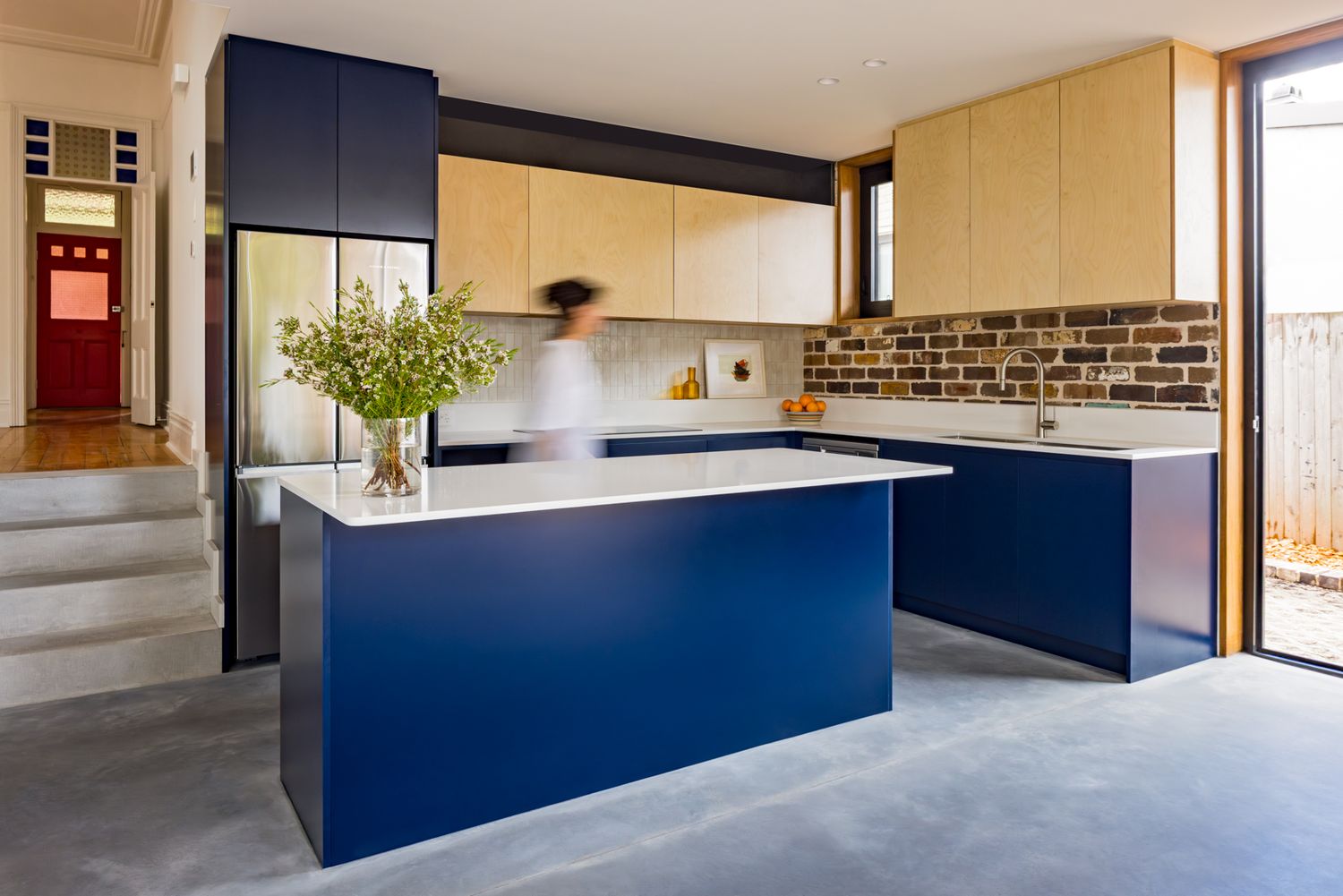 Contemporary kitchen with a navy blue island, white countertop, timber cabinets and an exposed brick splashback near a sunny window.