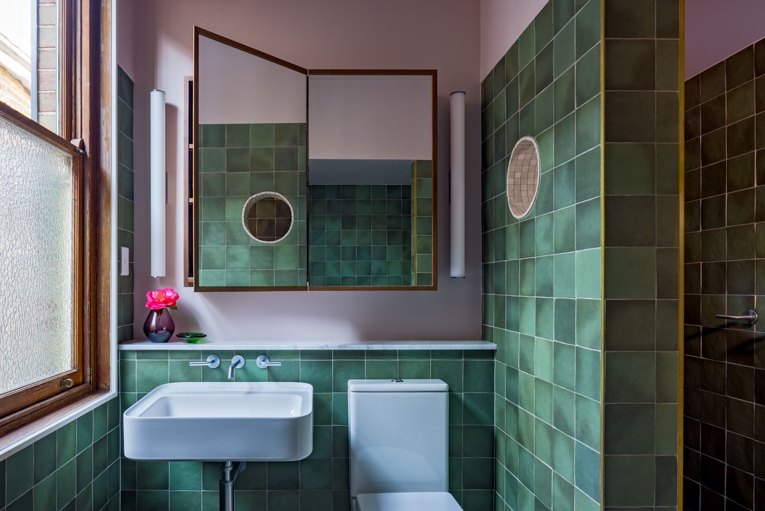 Small bathroom with glossy green tiles, a white wall-mounted basin, round mirror and a timber-framed window.