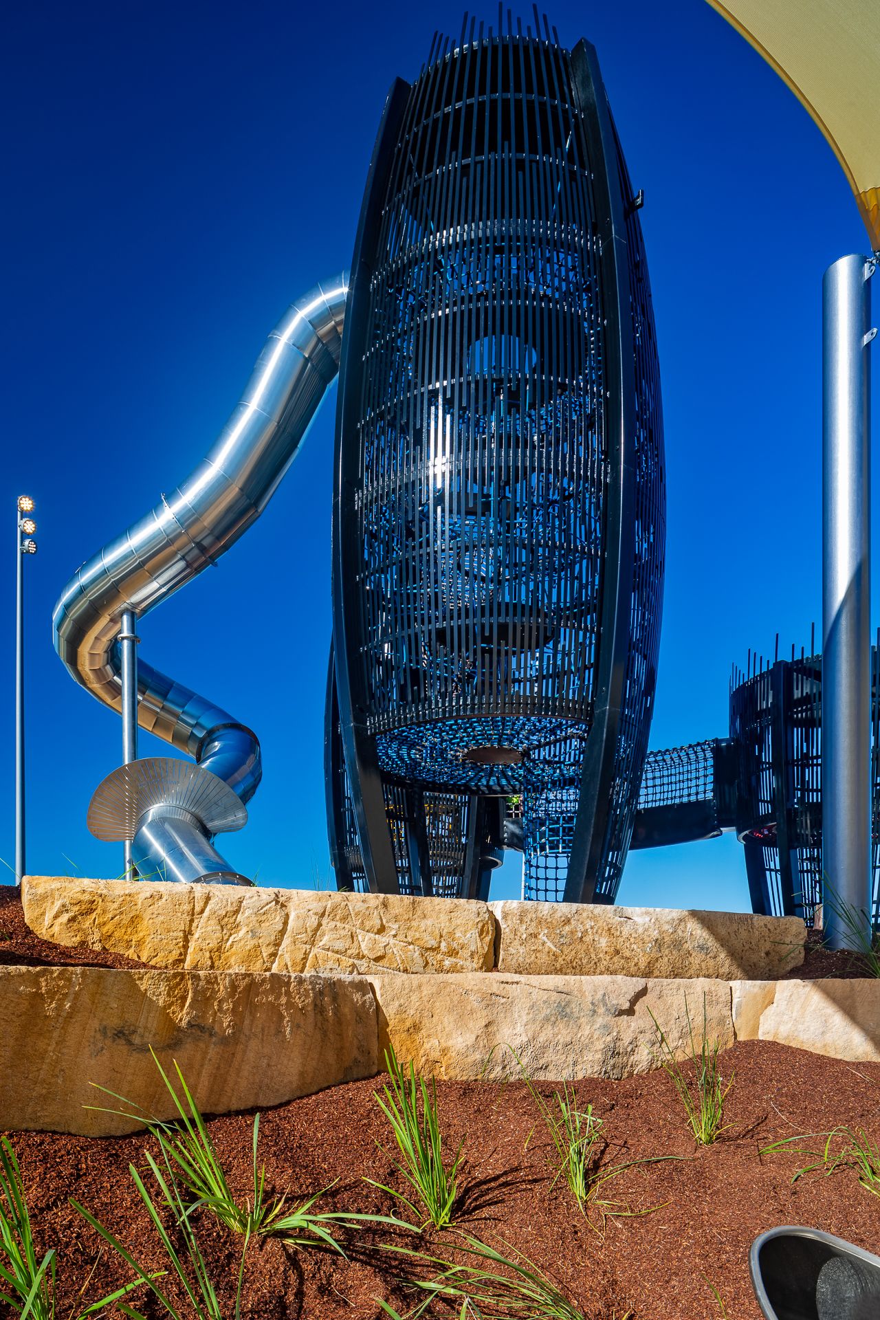 A giant black steel lattice tower with a curving silver slide rises above sandstone blocks and native grasses.