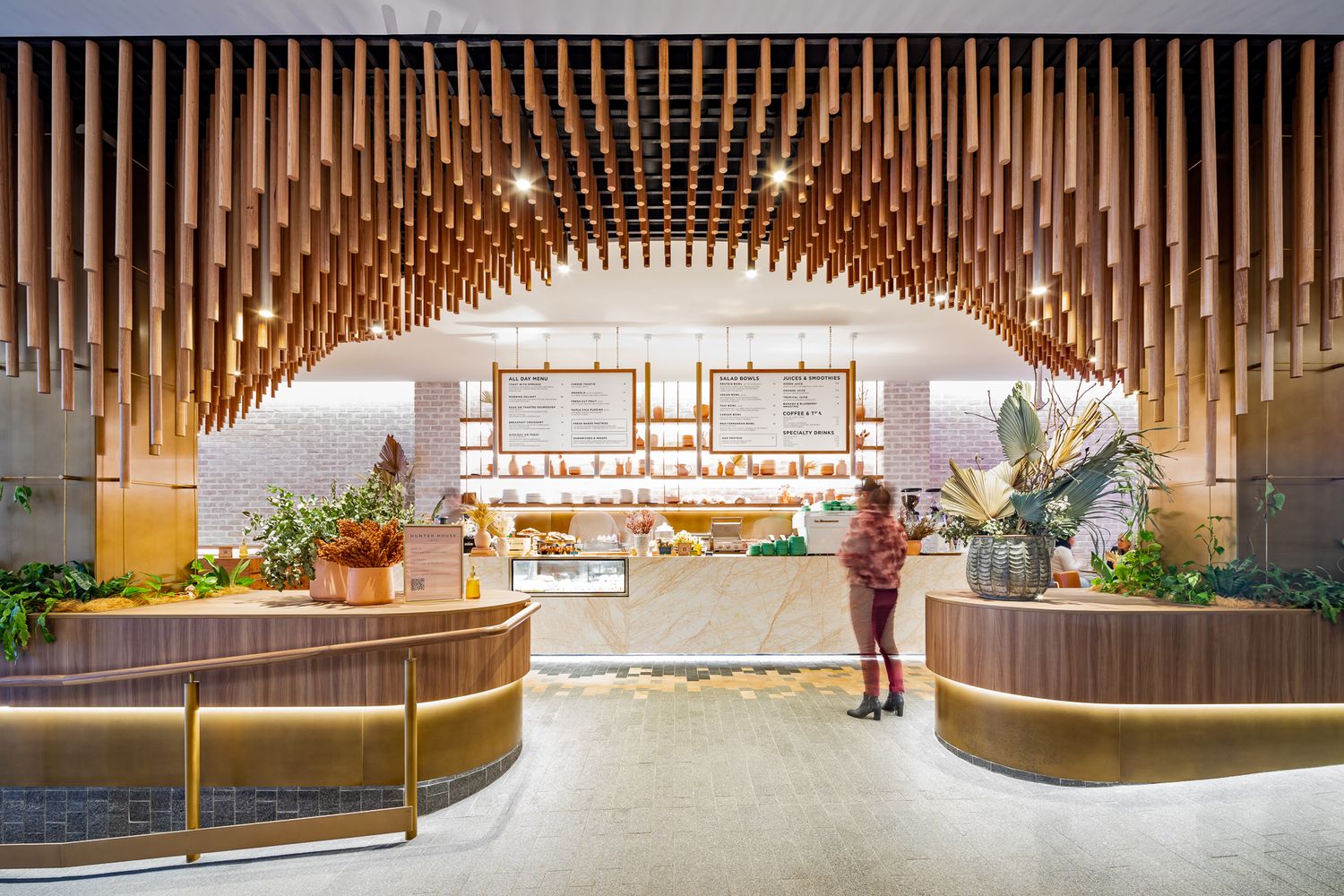 Visitor stands at a cafe entrance beneath an arched ceiling of vertical wooden slats, with curved timber planters and menu boards ahead.